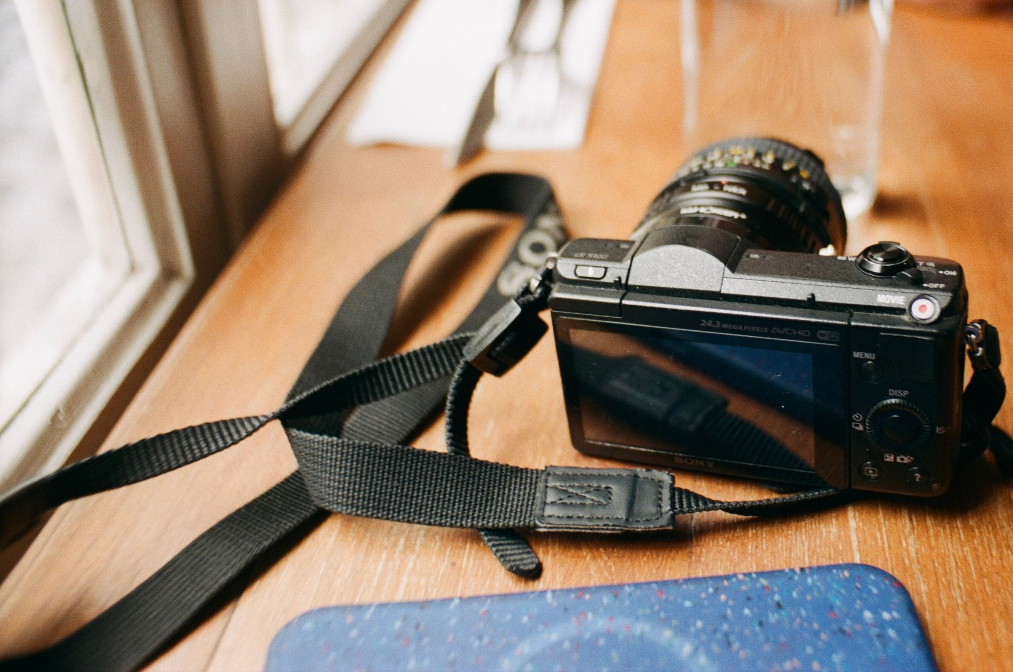 A table in a restaurant. A Sony mirrorless digital camera sits on it with a vintage prime lens attached with an adapter. An iPhone with a recycled plastic case is also visible.