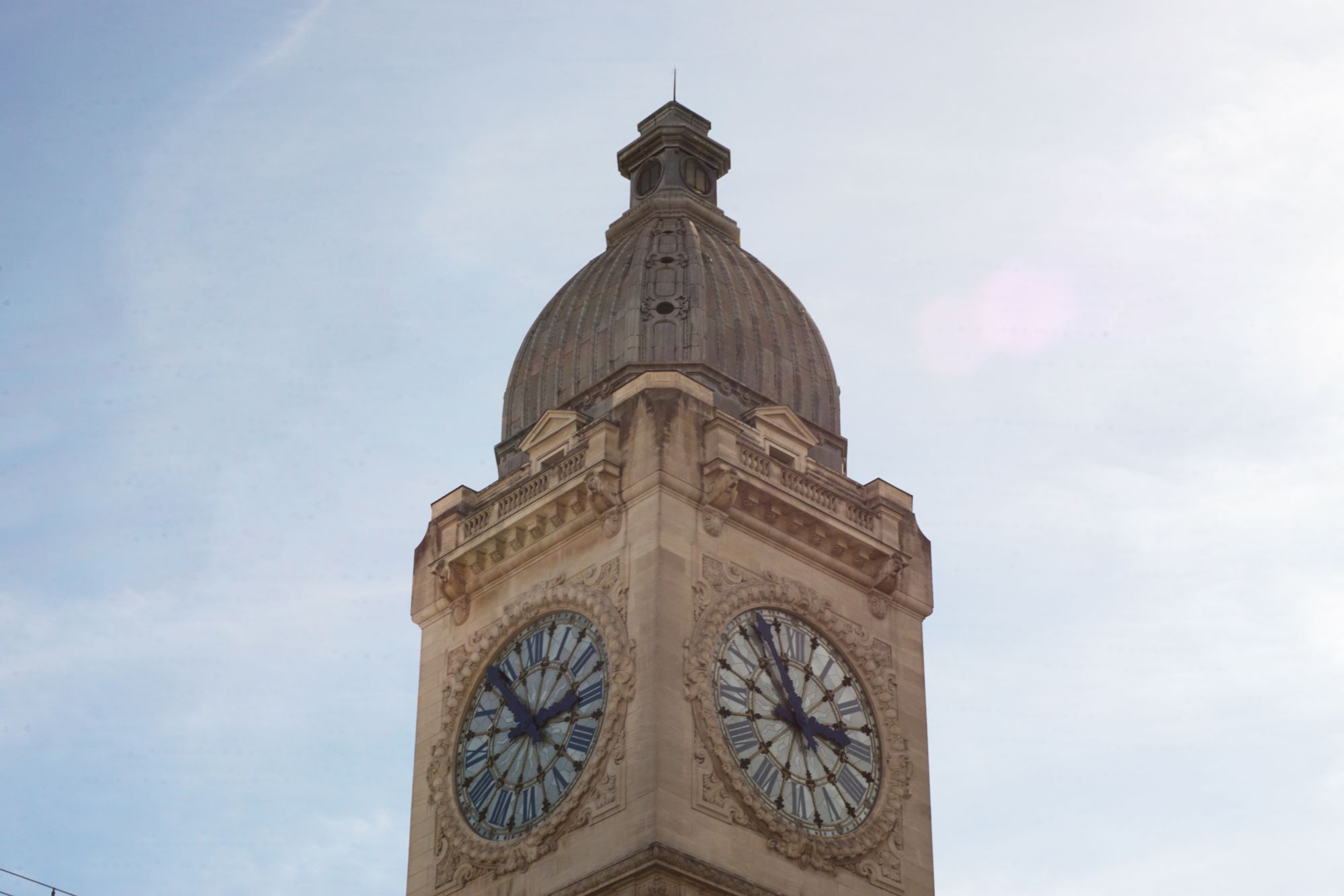 Telephoto shot of a limestone clock tower on a sunny day showing 2:55. The image is slightly streaked with small grey droplets where cleaning fluid hadn't dried on the sensor.