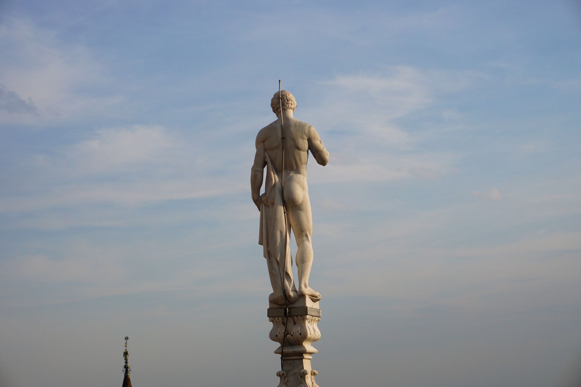 A nude male statue with short hair and carrying some cloth from behind, standing against a blue sky on a pillar. A lightning rod runs along the statue's back, passing between his buttocks.