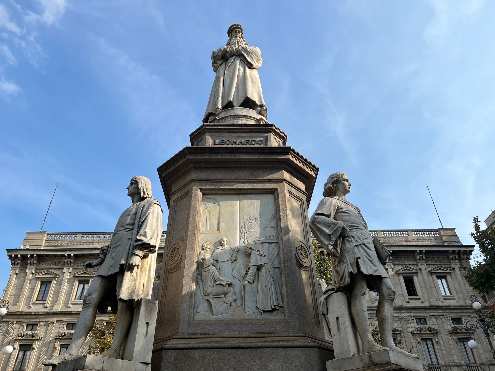 On a clear day, a statue of Leonardo da Vinci in a long robe, with men stood at all four corners and reliefs on each side of the plinth.