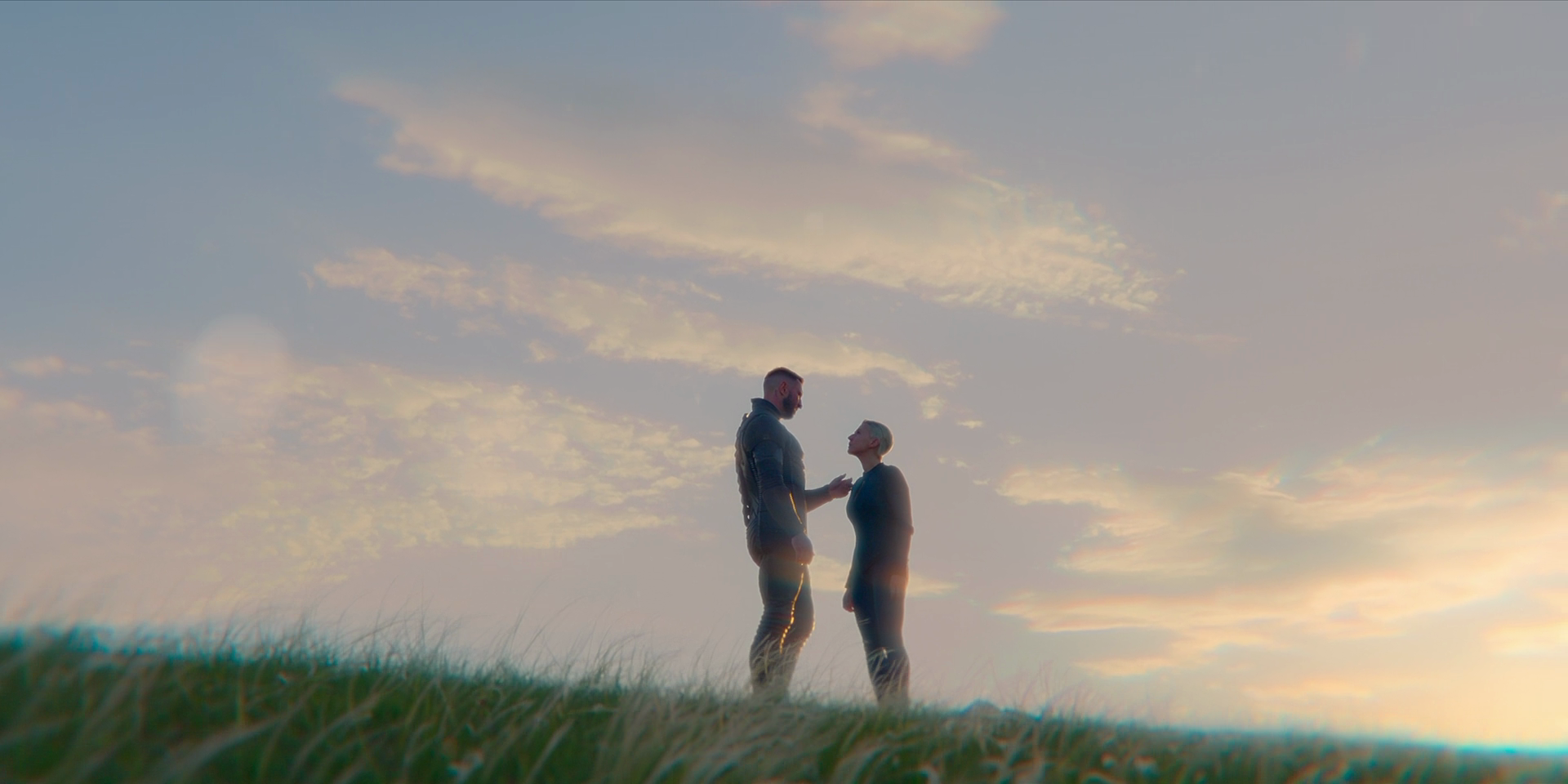 The Master Chief and Makee stand facing each other in a grassy field in dusk-like light. The Master Chief is moving his hand to place it on Makee's cheek.