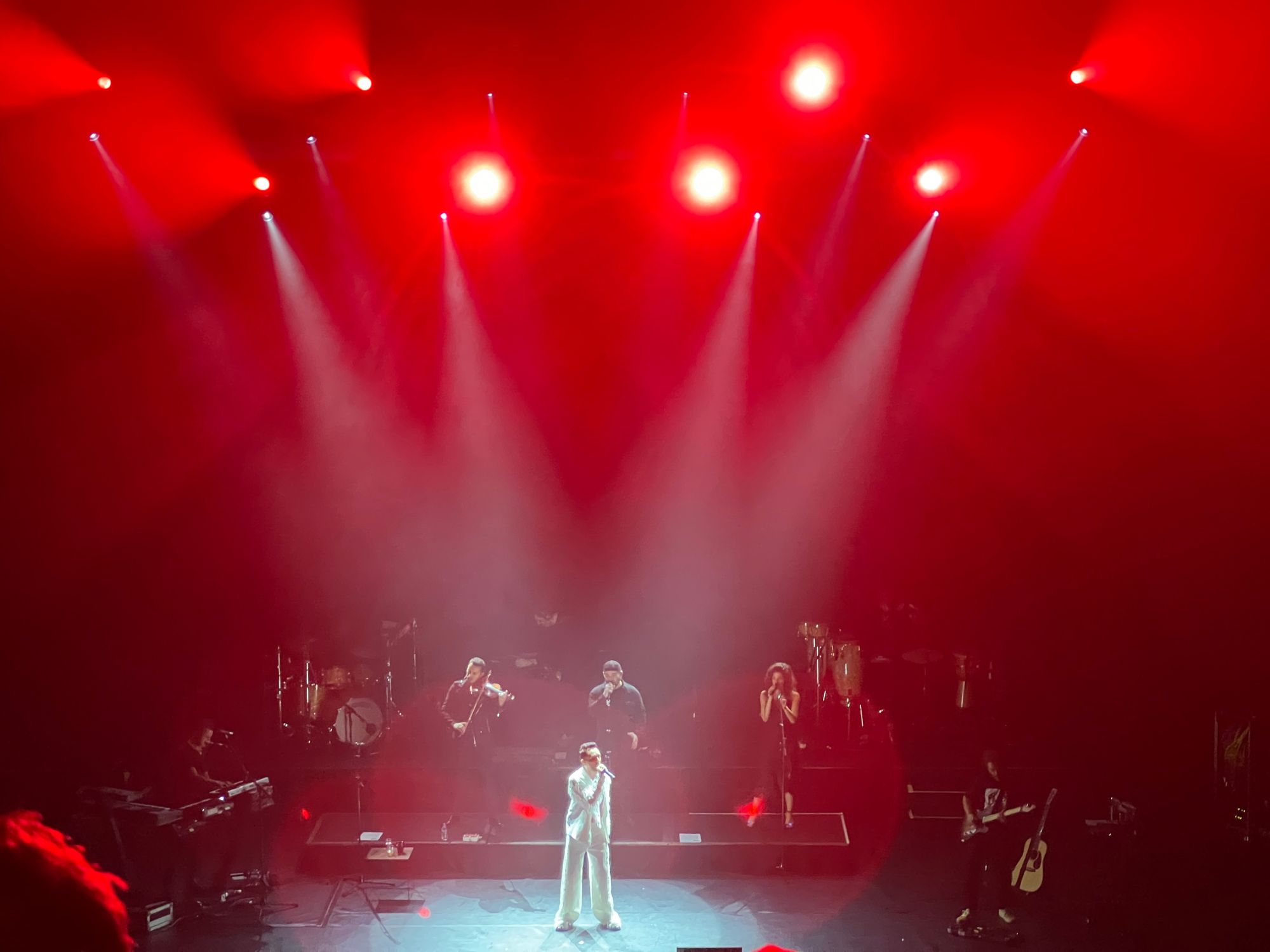 Mabel Matiz stands on a stage lit in red, wearing a bright white outfit, singing in front of his band who are all wearing black (two drummers, keyboardist, violinist, two backing singers, and violinist.)