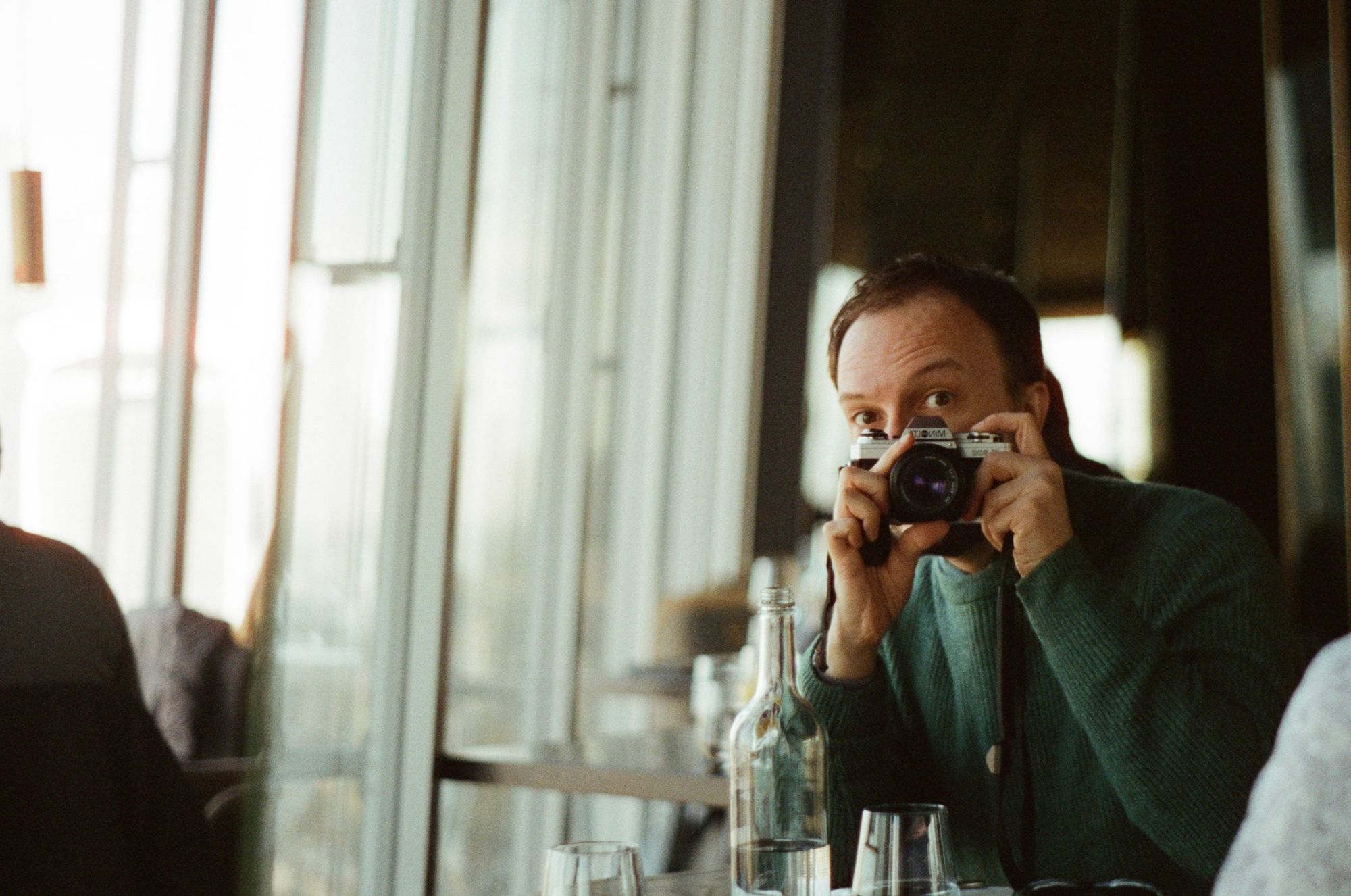 Jonathan sits at a table in a restaurant with floor-to-ceiling windows, photographing himself with an old Minolta SLR camera in a mirror. He's wearing a green jumper.