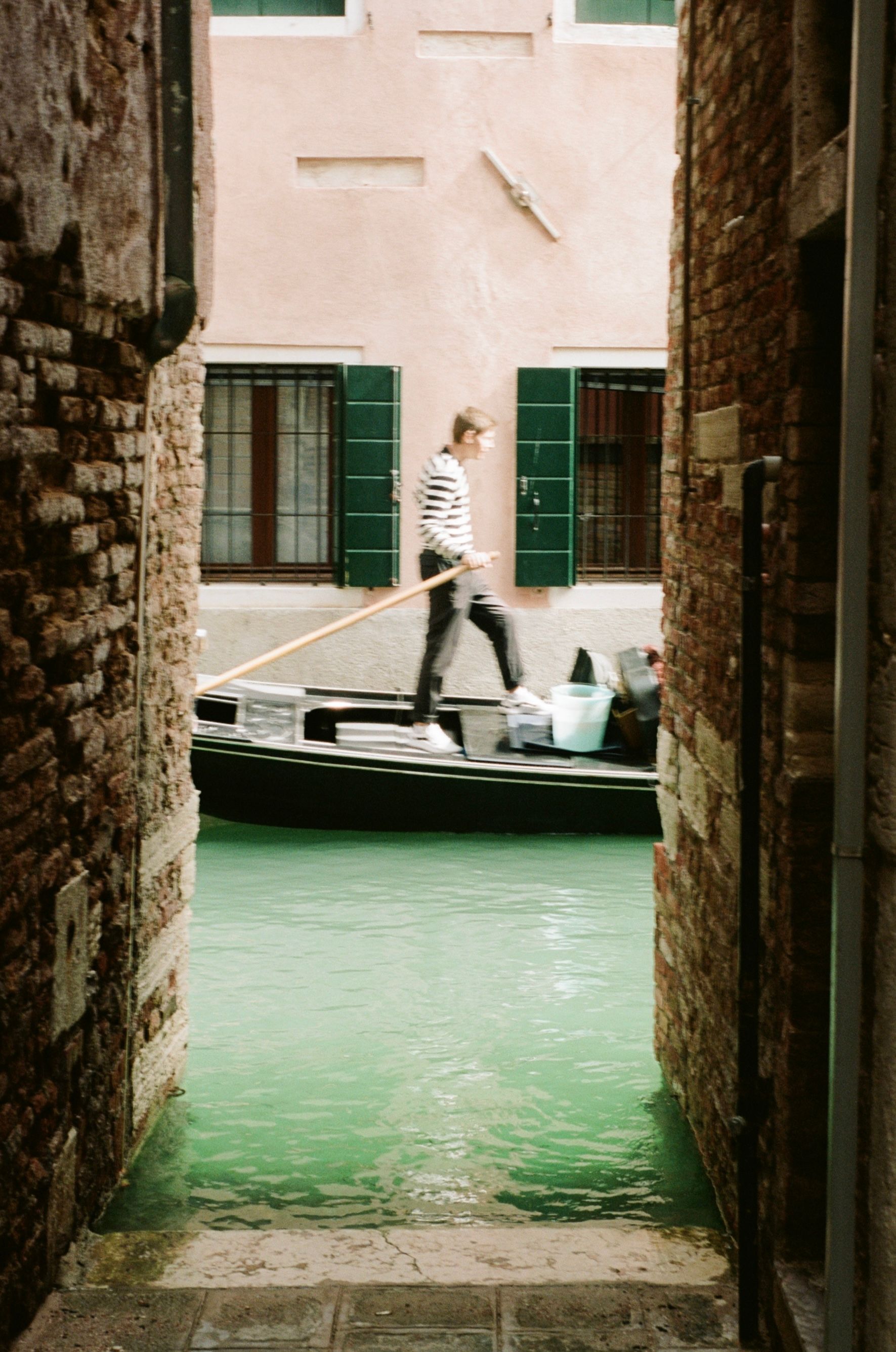 Down an alley that terminates in a canal with the water almost flooding the pavement, green shutters on the walls opposite; a young gondolier, male, lithe, with brown hair and in the iconic white-and-dark-blue stripy jersey, stands on his vessel, oar comfortably pointing aft with his right hand resting on it. He shoots off to the right with ease.