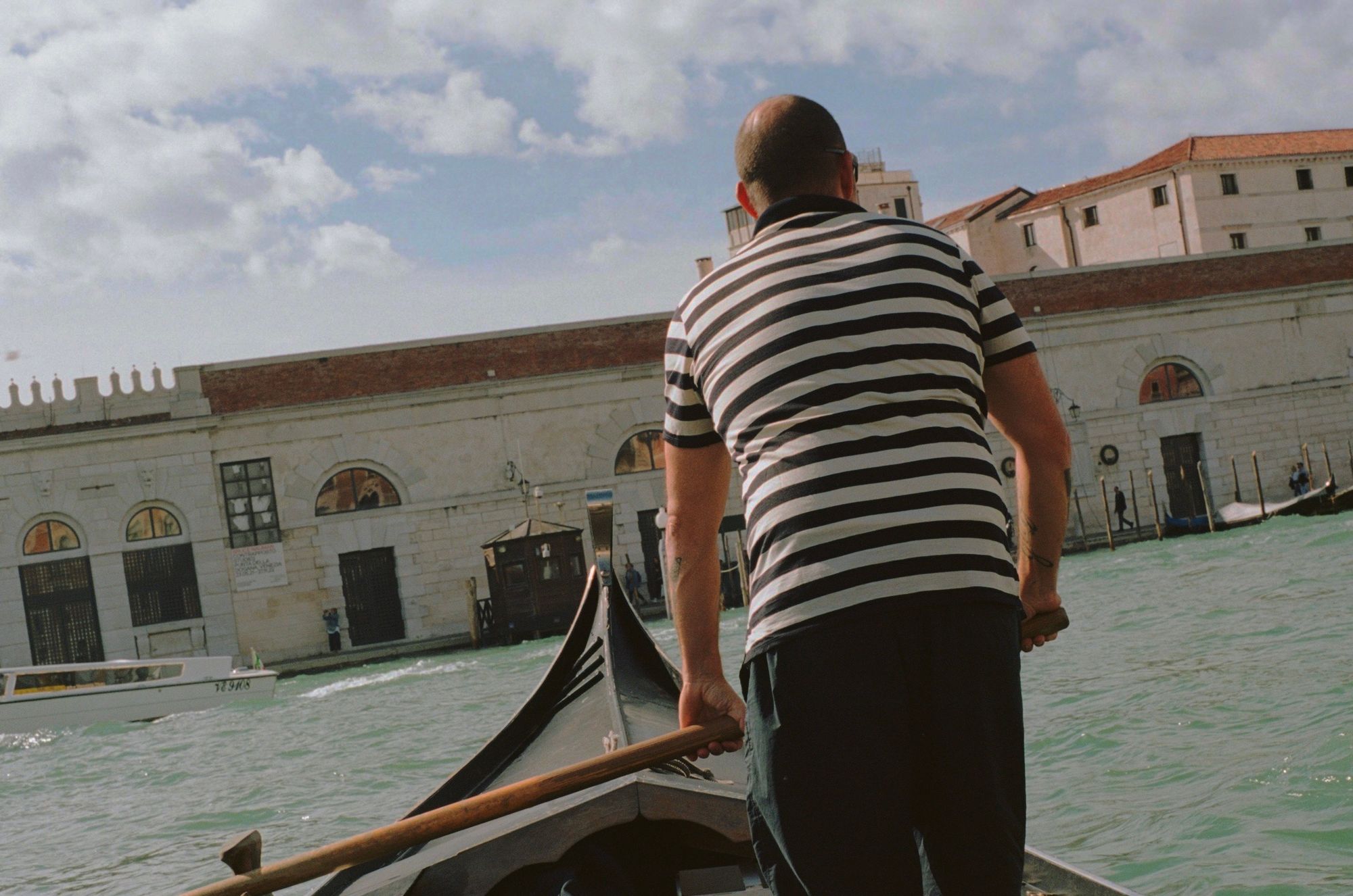 A heavy-set, middle-aged gondolier with cropped hair and a tattoo, seen from behind, working as a ferryman at the front of a large gondola—he looks to the right as he approaches Salute, making adjustments with his oar.