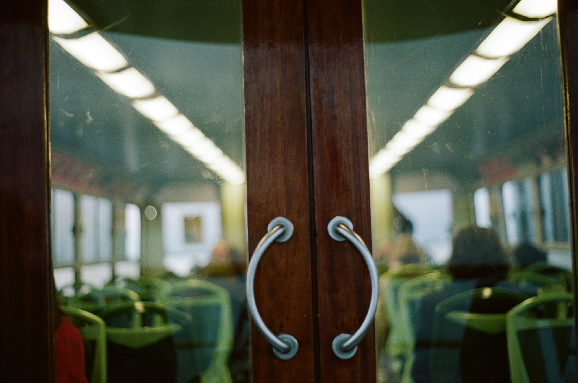 Wooden doors with glass windows on a vaporetto looking inwards at the seating from behind, about half occupied. Everything has a pleasing warm dusky glow.