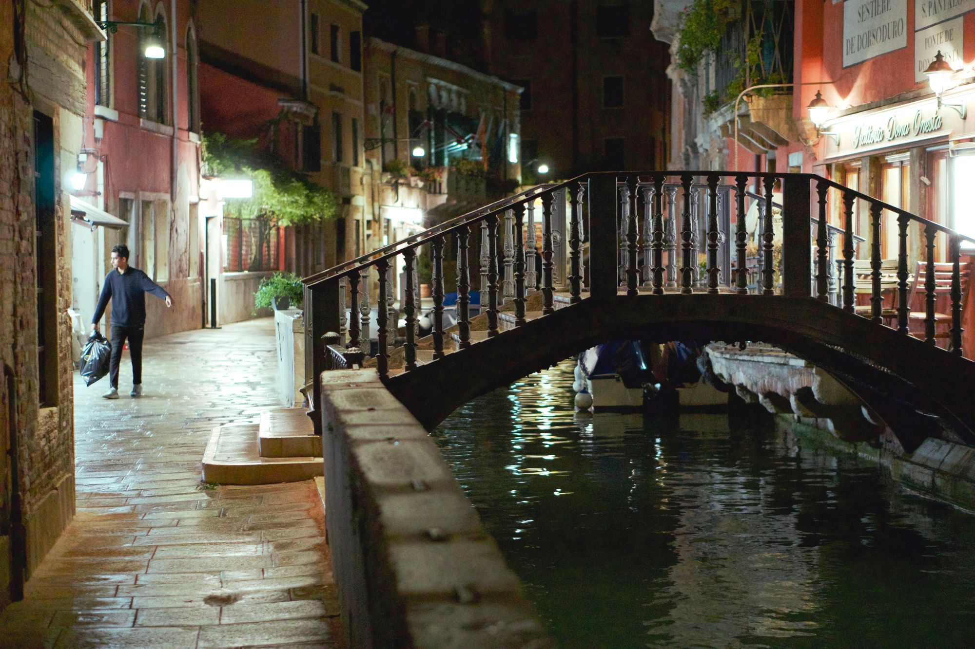 An arched stepped footbridge over a narrow canal at night (Ponte de Dona Onesta) with lights on the shops. A man in a black jumper and black trousers carries out a black bin bag for disposal.