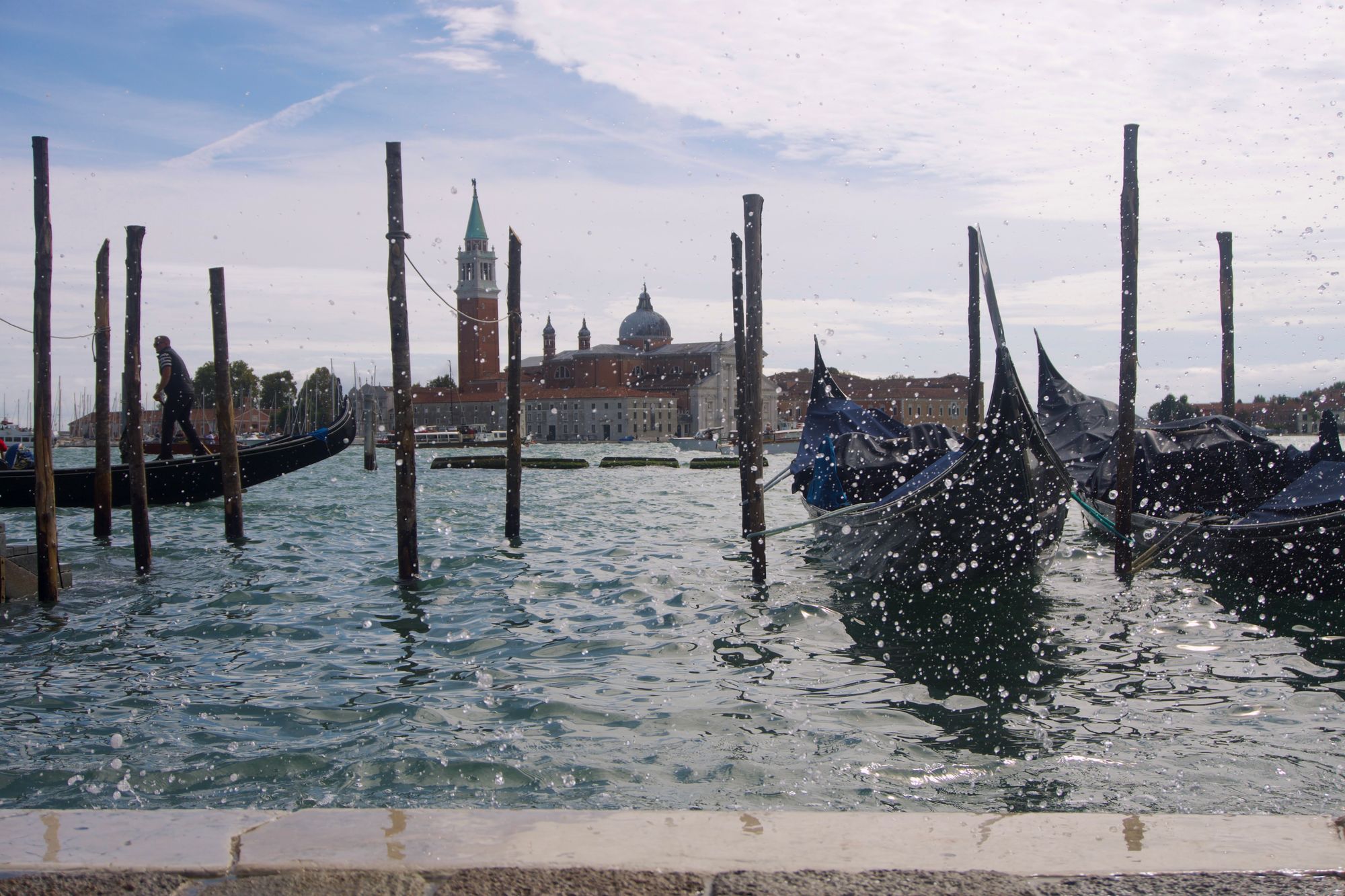 Gondolas moored against long wooden poles bob up and down in the Venetian lagoon, which whips up in spray before us as we stand perilously close to the edge (the turquoise waters almost spilling over.) In the background, we see San Giorgio Maggiore's dome and tower, obstinate.