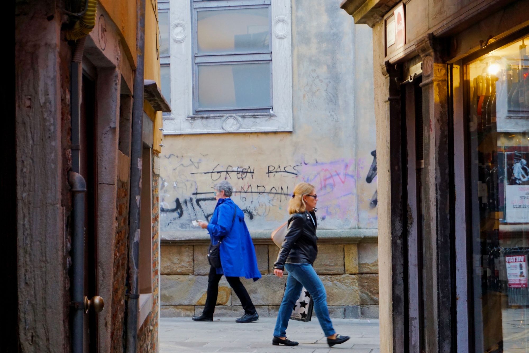 To the end of an alley between two buildings at sunset, against a wall with graffiti (reading "GREEN PASS TAMPON" which has been crossed out): a woman in a leather jacket with shades, and a man in a blue raincoat, walk in opposite directions.