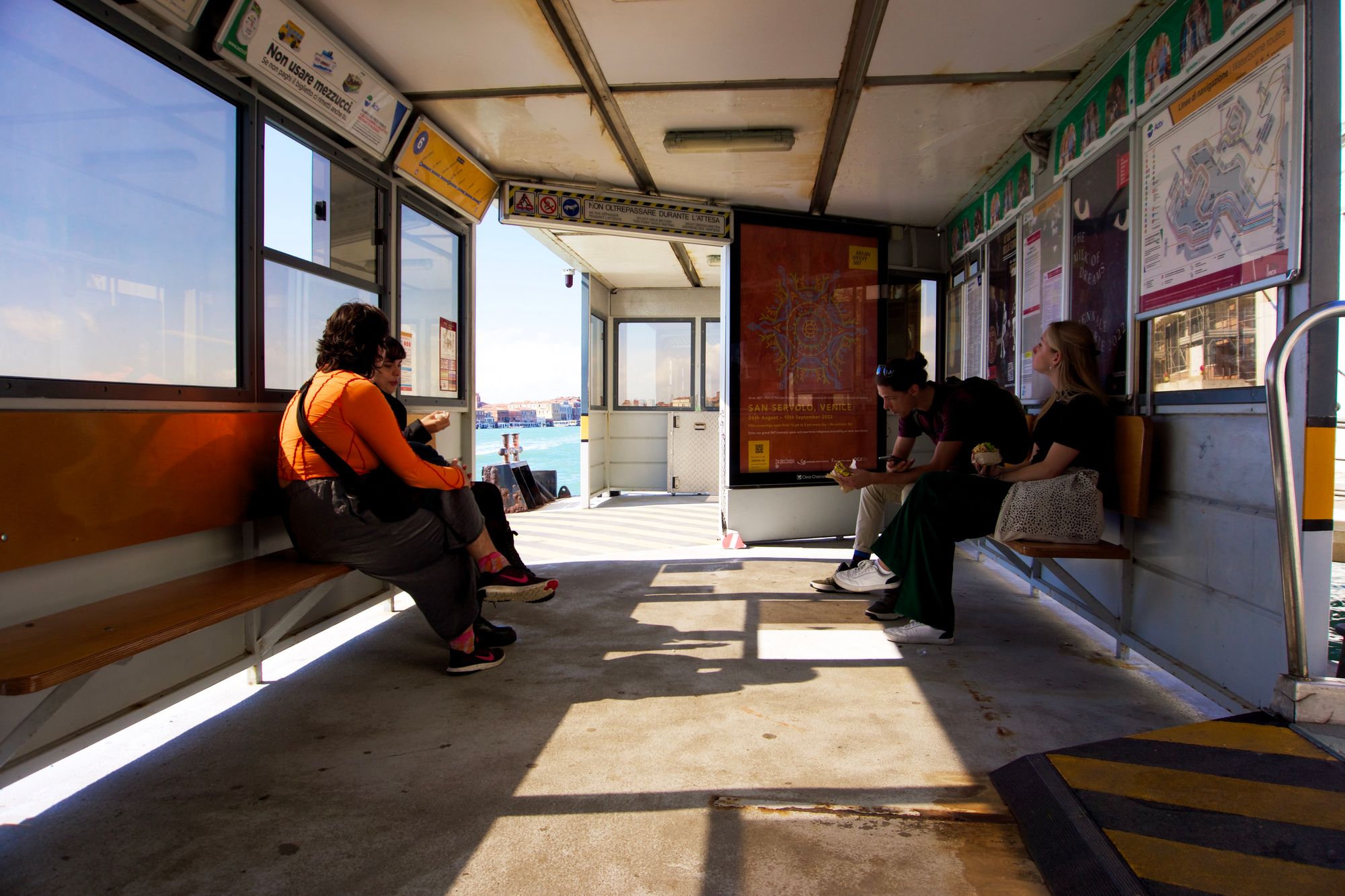 On a vaporetto pier on a sunny day with light shining shadows through the windows. Four other people sit on benches looking at their phones our out of the open boarding area.