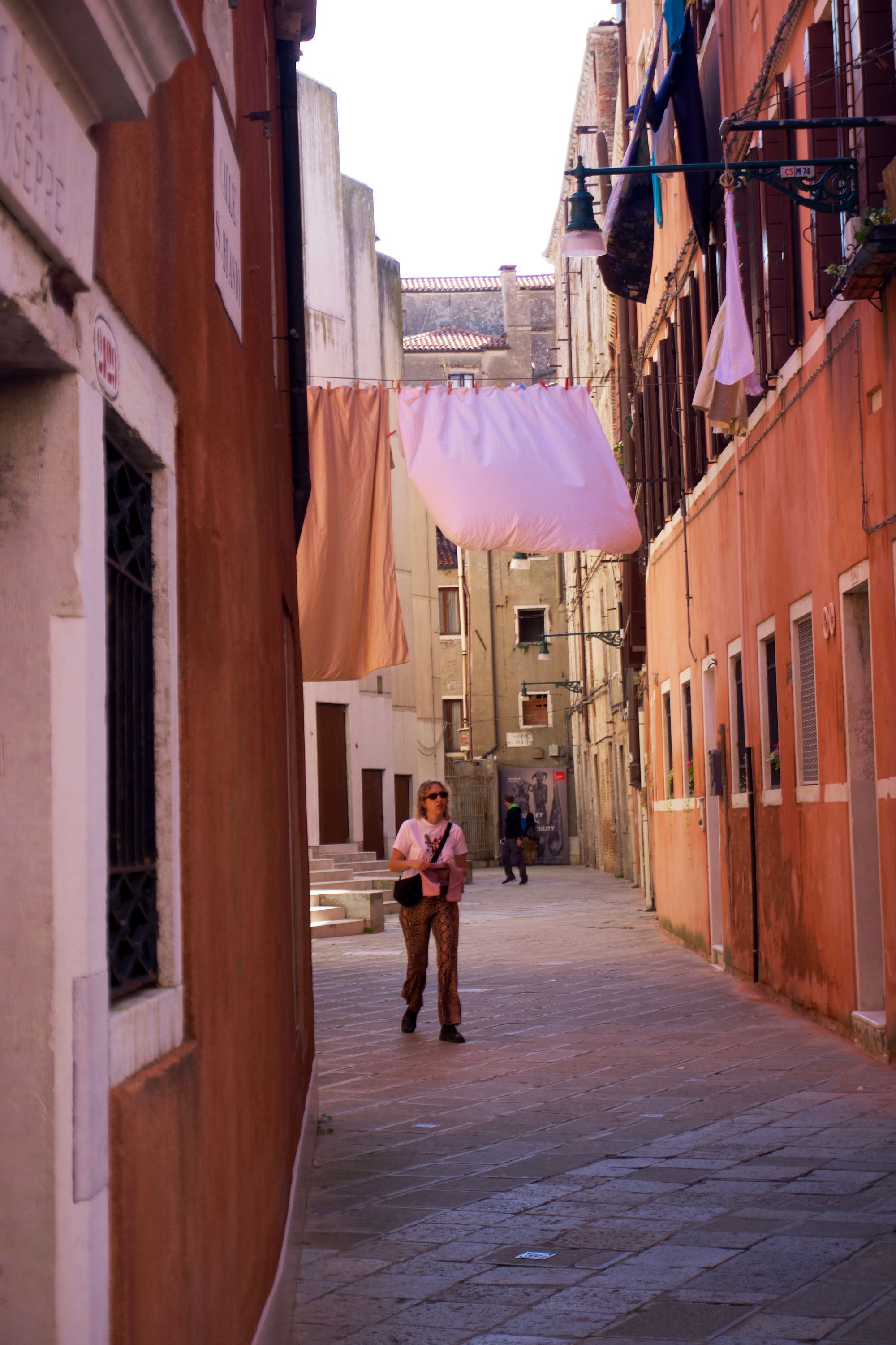 A person in a grey t-shirt and brown flared trousers walks down a narrow pedestrian street with terracotta-coloured houses on either side. Above their head, a washing line hangs, with a brown and pink bedsheet flying from them attached with red pegs.