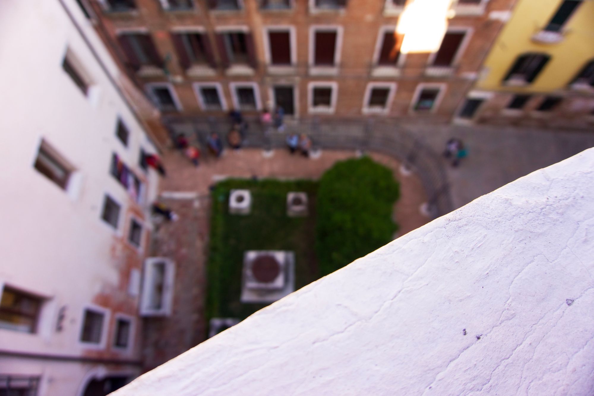 View down on a small garden on a terrazzo square, with people queuing outside the fence. It's inside a courtyard formed by several larger buildings in white, terracotta, and yellow. A reflection casts a sharp white rectangle on the terracotta building. In the foreground, a balustrade is painted white in sloppy ridges.
