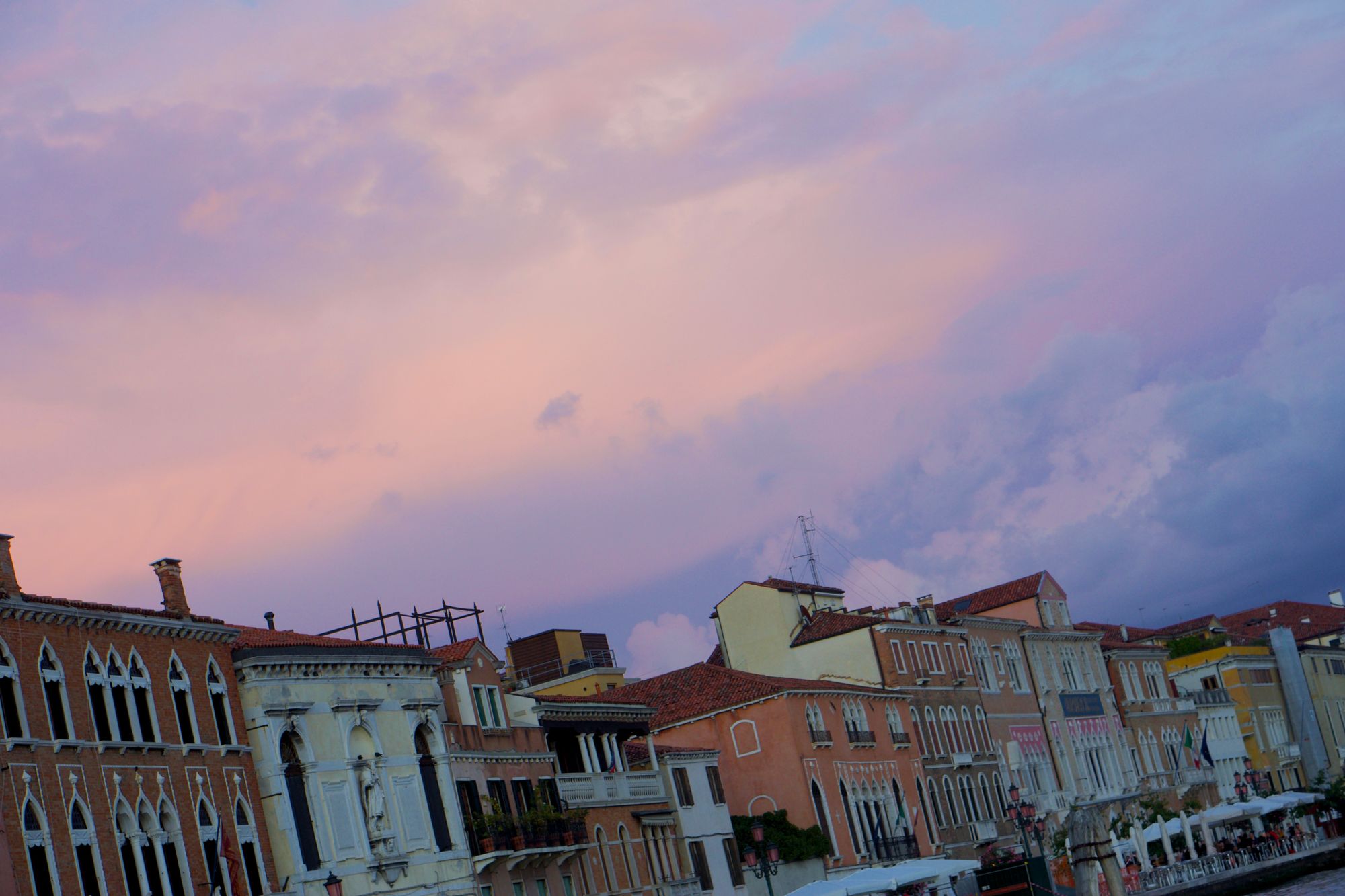 A parade of waterfront buildings with fluffy blue-pink-salmon coloured clouds behind them.