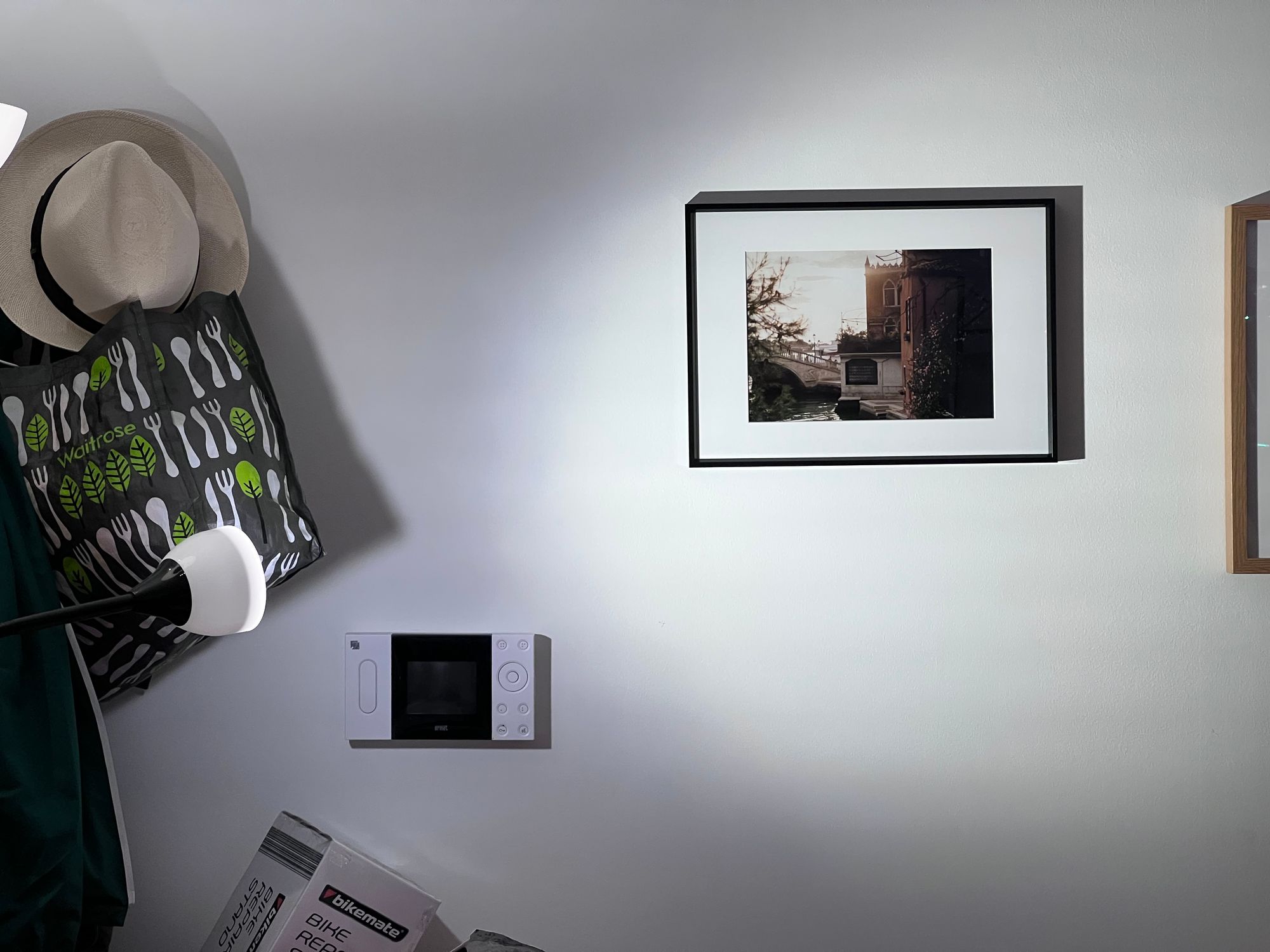 A white wall in a flat with a door intercom, the top of a box containing a bike repair stand, a straw hat and a bag for life hanging from coat hooks. In a black frame with a white mounting, a photo in the golden hour from Venice, of a bridge over a canal with a tree on one side and a creeping plant with pink flowers sticking to the terracotta coloured wall of the building on the right.