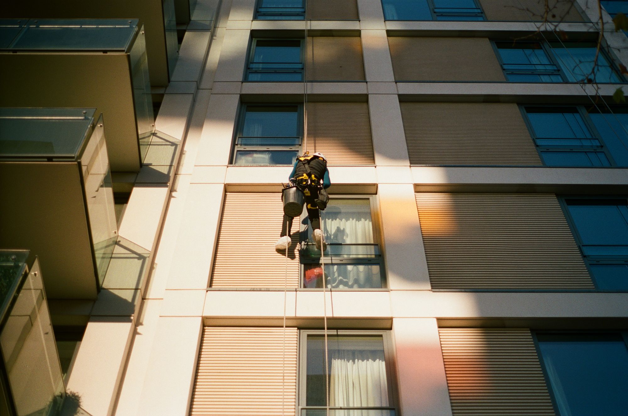 A window cleaner abseiling down the side of a block of flats with wooden slat cladding in a pool of light cast through glass balcony balustrades. He carries a bucket, wears a white helmet, a black gilet, and white oversells over his shoes.
