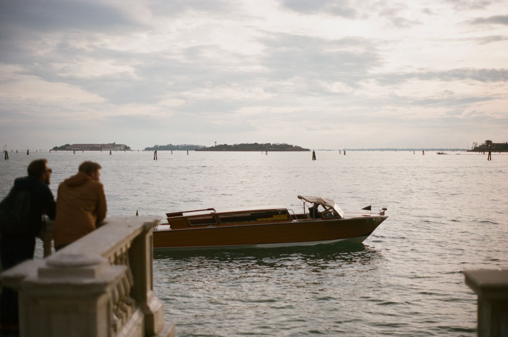 Two men lean on a balustrade watching out over the lagoon at the many islands and wooden buoys, as a water taxi zips past, in a warm pastel-like evening light.