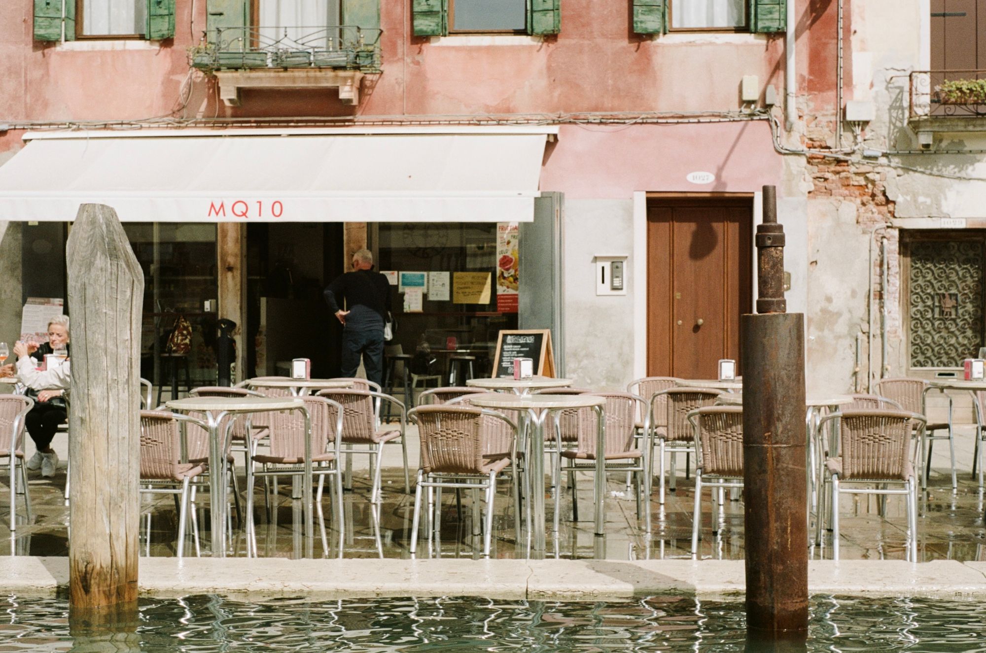 A café alongside a canal with white awning reading "MQ10" in red text. The water is dangerously close to overspilling the canal wall, and some already has, wetting the pavement beneath the al fresco seating.