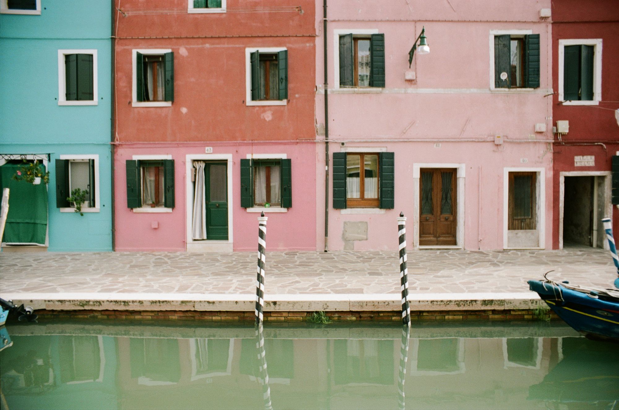 A row of colourful terraced houses (sky blue, dark pink, pale link, burgundy) with green window shutters open out onto a canal towpath with light-coloured crazy paving. The canal itself is aquamarine, reflecting the houses, and two black-and-white spiral-striped mooring posts. A small blue boat's prow sticks into the frame from the right.
