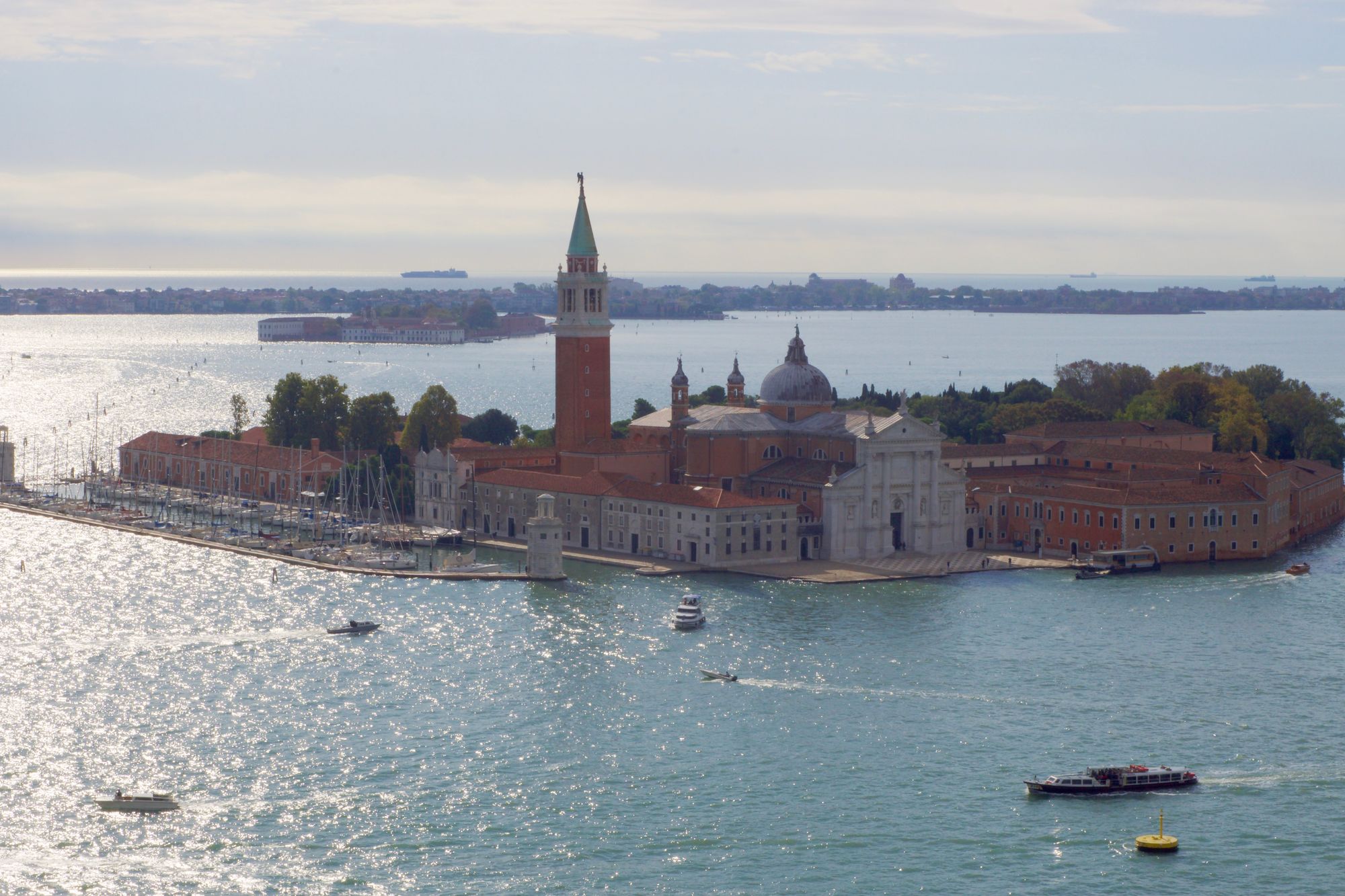 The island of San Giorgio Maggiore seen from a tower on a nearby island, the lagoon glimmering in the morning light as boats zip past, the terracotta of the church obstinate in the hazy light.