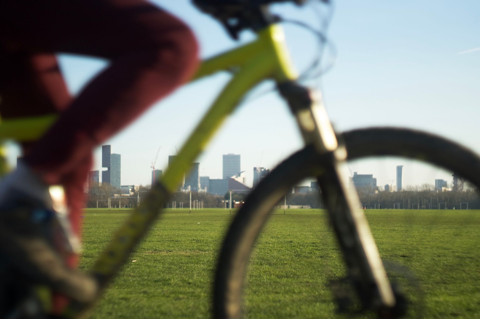 A shot over a green field towards a skyline on a sunny day. In the foreground, a person with red trousers on a neon-green mountain bike crosses the frame.