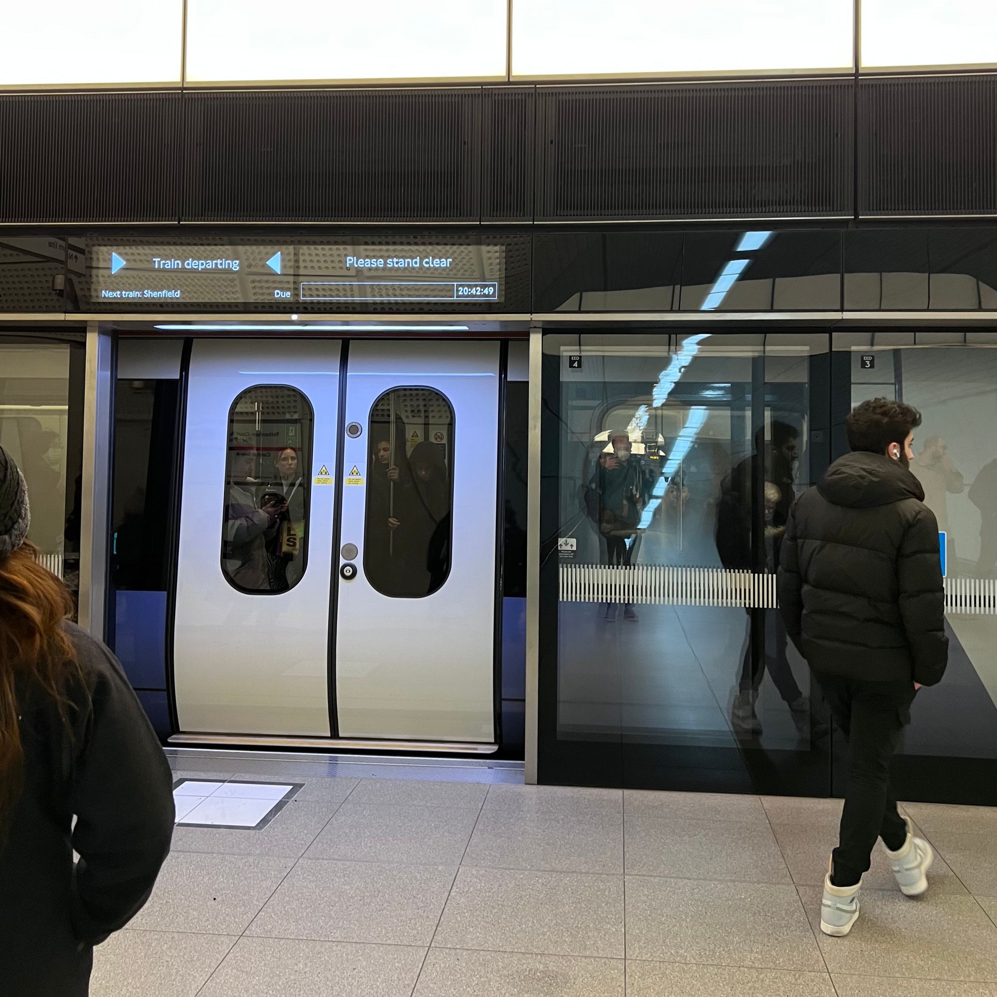 An Elizabeth line train stood at a platform with its doors closed, but the full-height platform edge doors still open. The screen displays "Train departing, please stand clear," but the train is not departing.