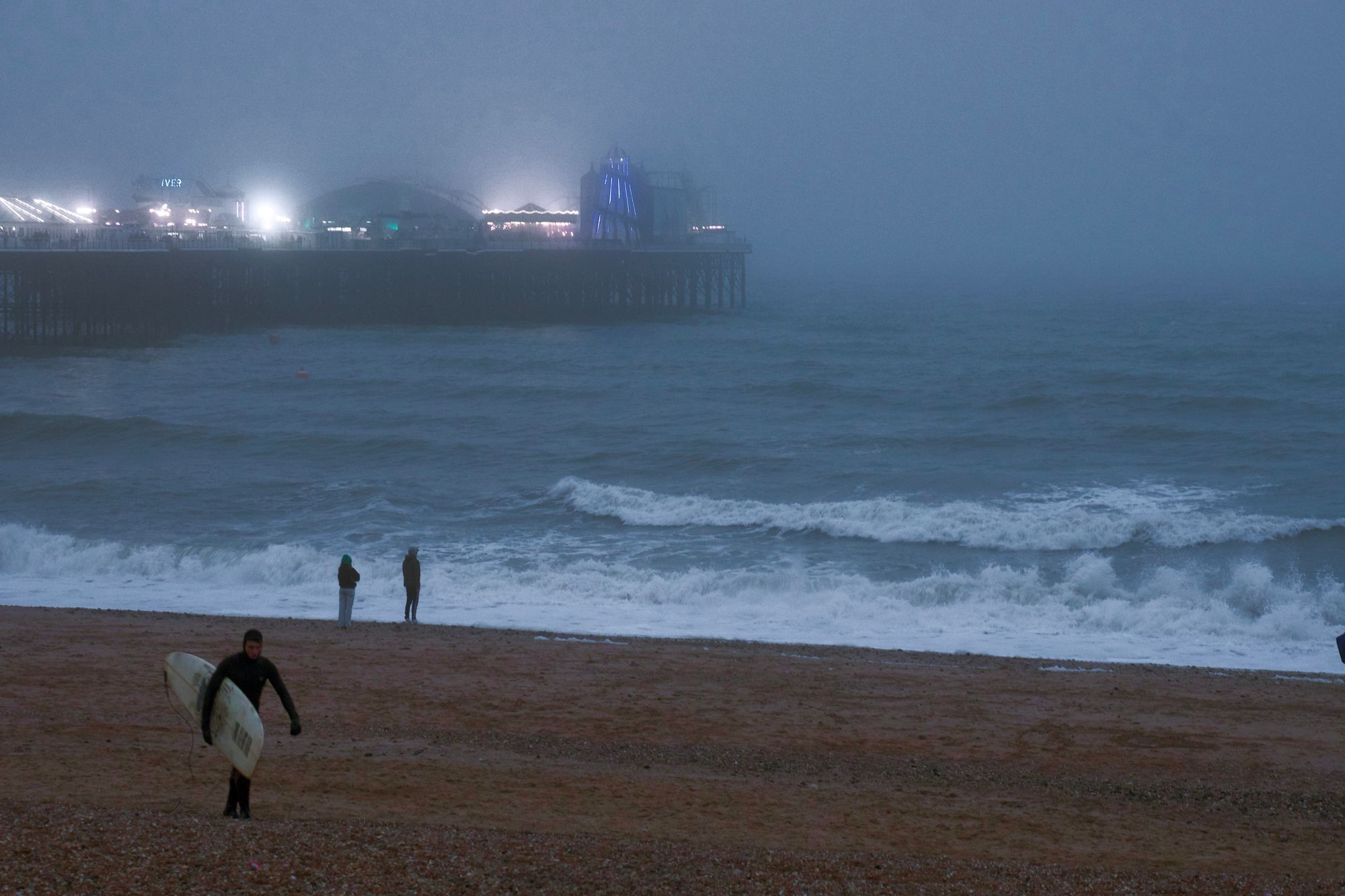 A choppy sea on a grey day, with Brighton Pier in the background. Two people stand on the brown shingle beach looking out to sea. A man in a wetsuit carrying a surfboard walks up the beach, looking astonishingly chilly.
