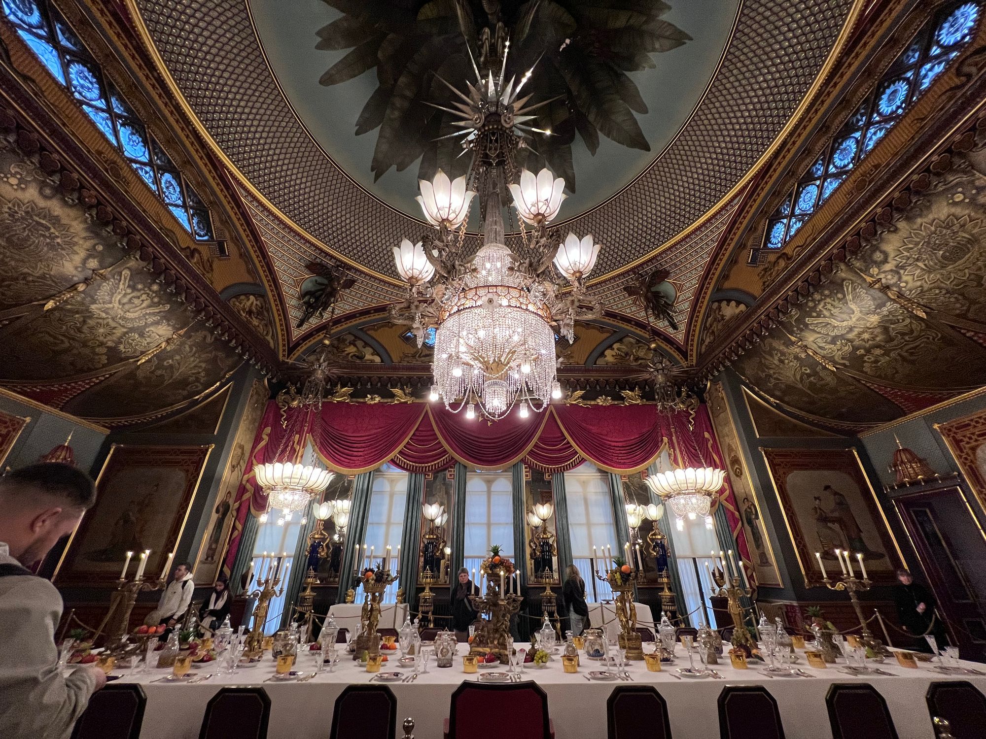 A large dining room with a domed ceiling (from which hands the chandelier in the header image). There are additional chandeliers at each corner, red gilded curtains and drapes, and every border and crevice is gilded in some way. The table is long and covered in a white tablecloth, laid out as if for food. Visitors surround the table.