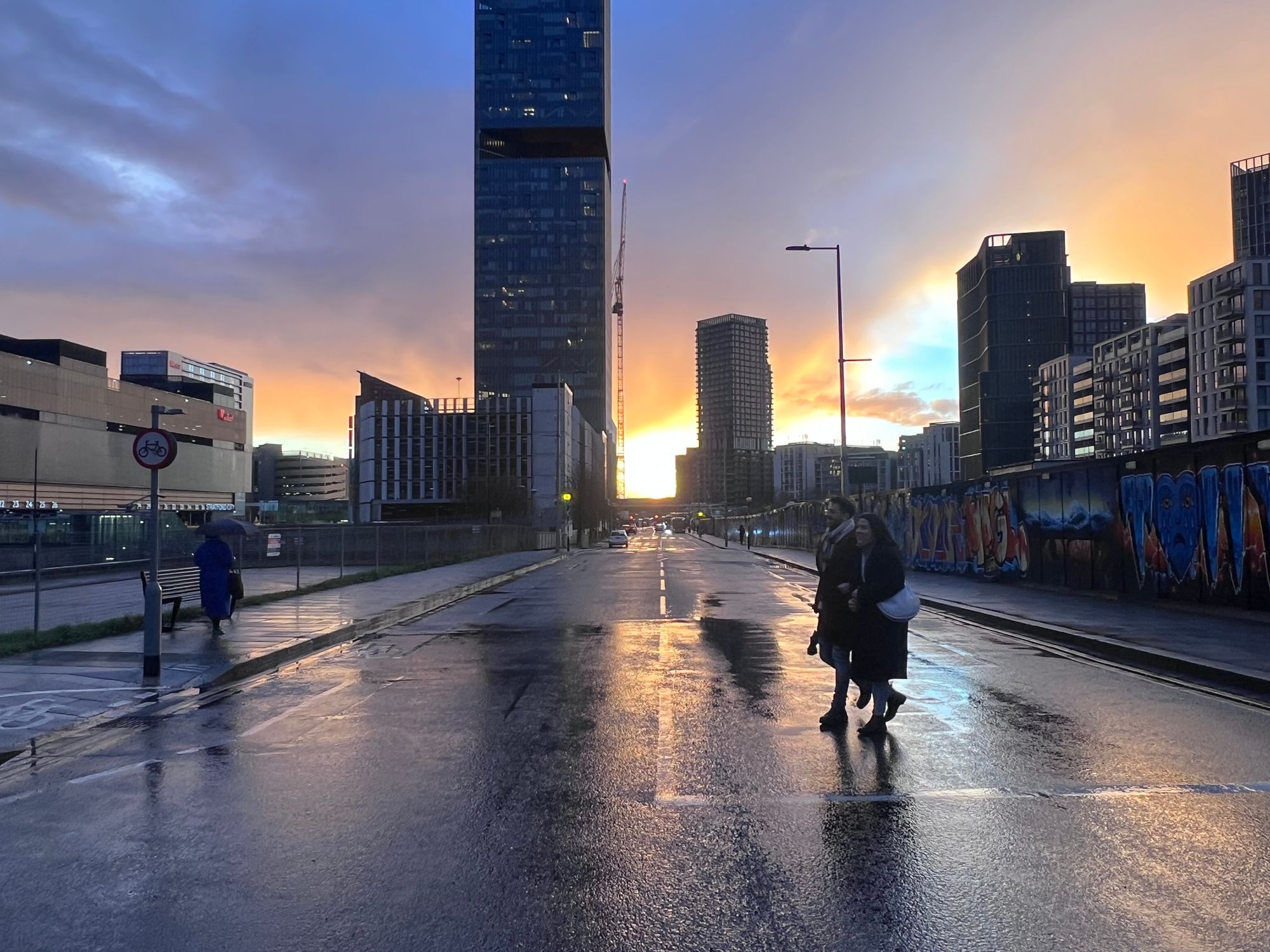 People cross a wide street with tall buildings in the distance, the tarmac wet and reflecting a glorious cloudy sunset.