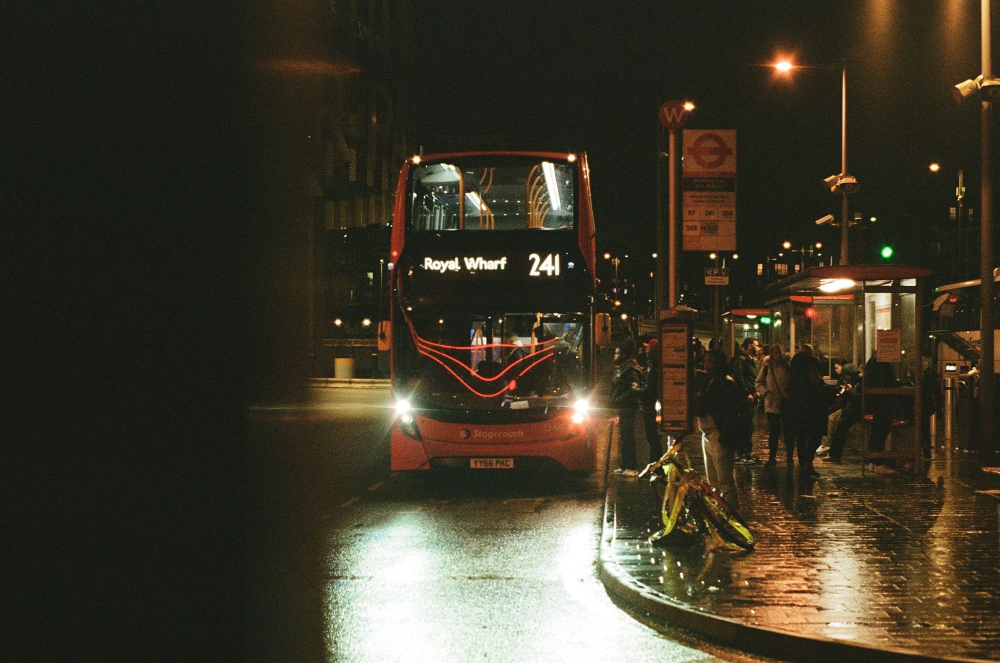 A rainy bus station. A red double-decker bus (the 241 for Royal Wharf) is boarding. Three neon strip lights reflect into the windscreen.