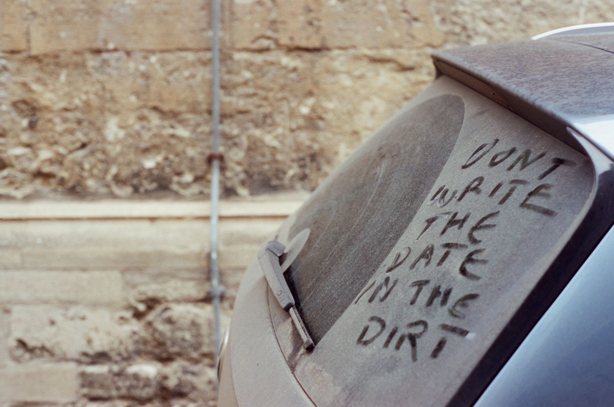 The back of a car window covered in dust, with the words "DONT WRITE THE DATE IN THE DIRT" written in the dirt. Behind it is an old stone-brick wall.