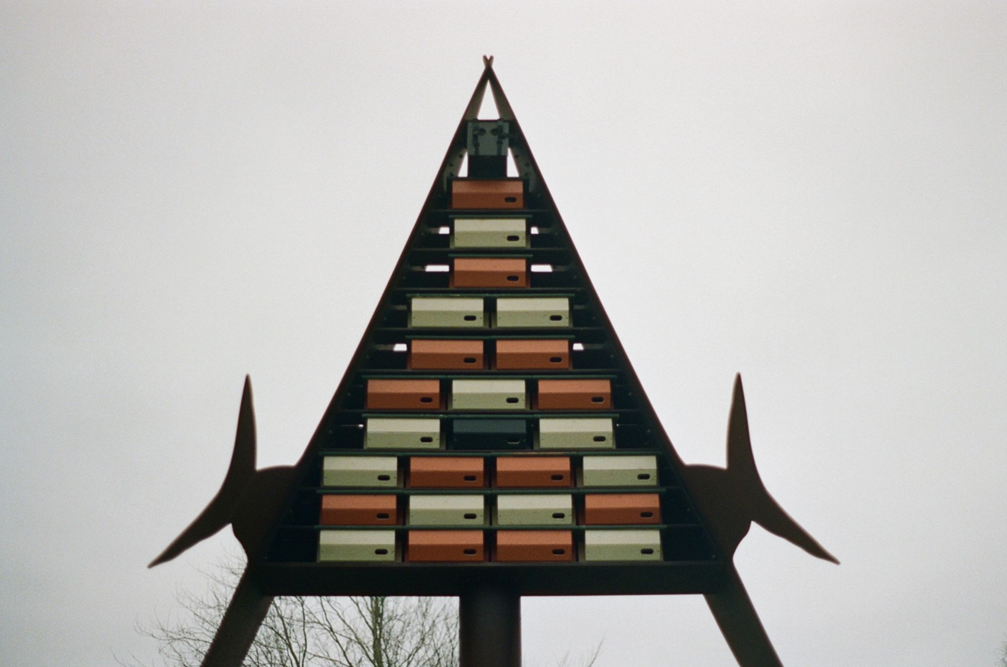 A large triangular structure on decorative metal legs with protrusions that look like swallowtails. Inside are stacked horizontal boxes with a hole in each, in orange, beige and black—these are nesting boxes for swifts.