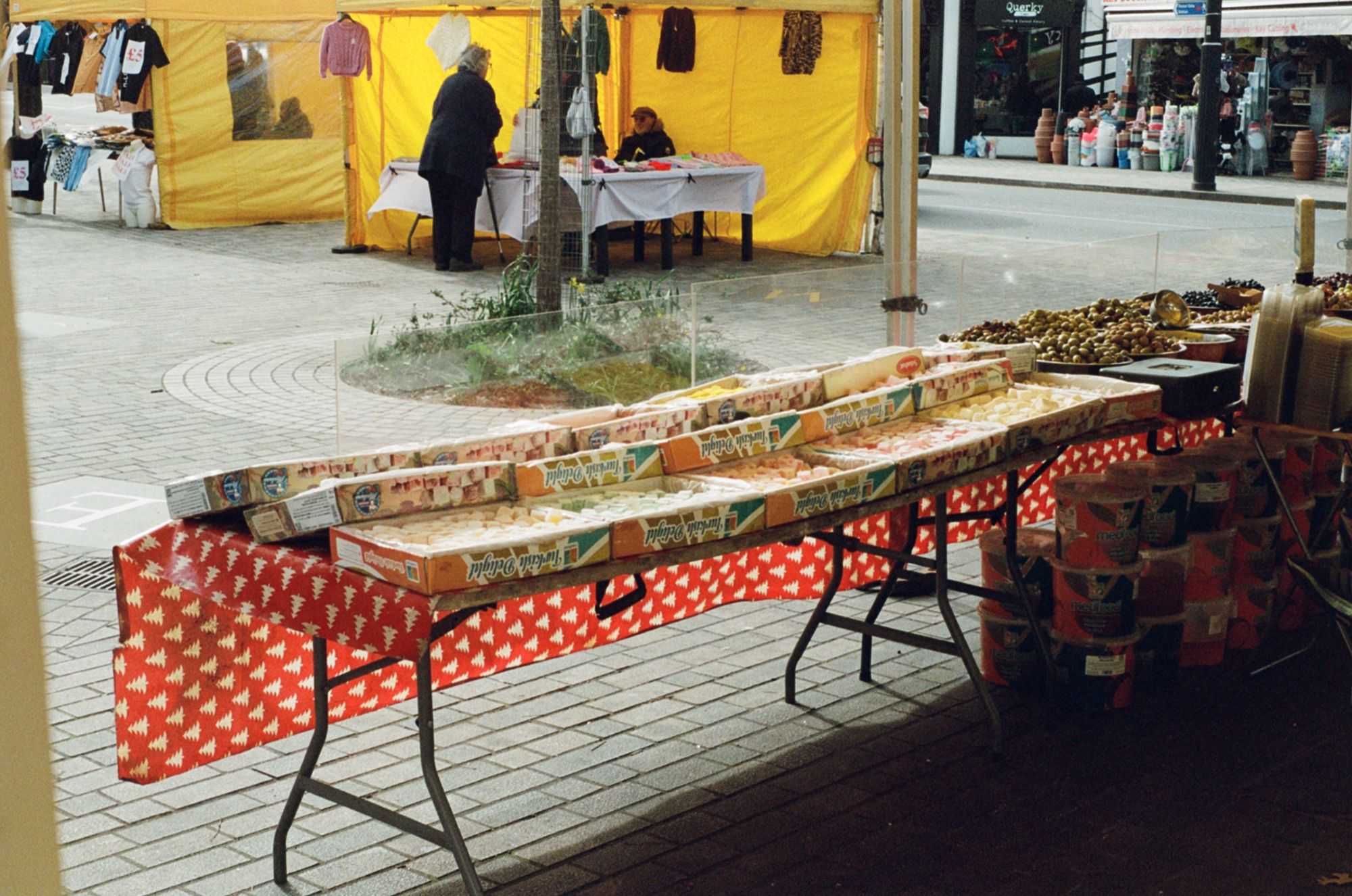 A market stall with a long table with red tablecloth, seen from behind. Lots of open boxes of Turkish delight/lokum in various colours line the table, along with olives and various other produce.