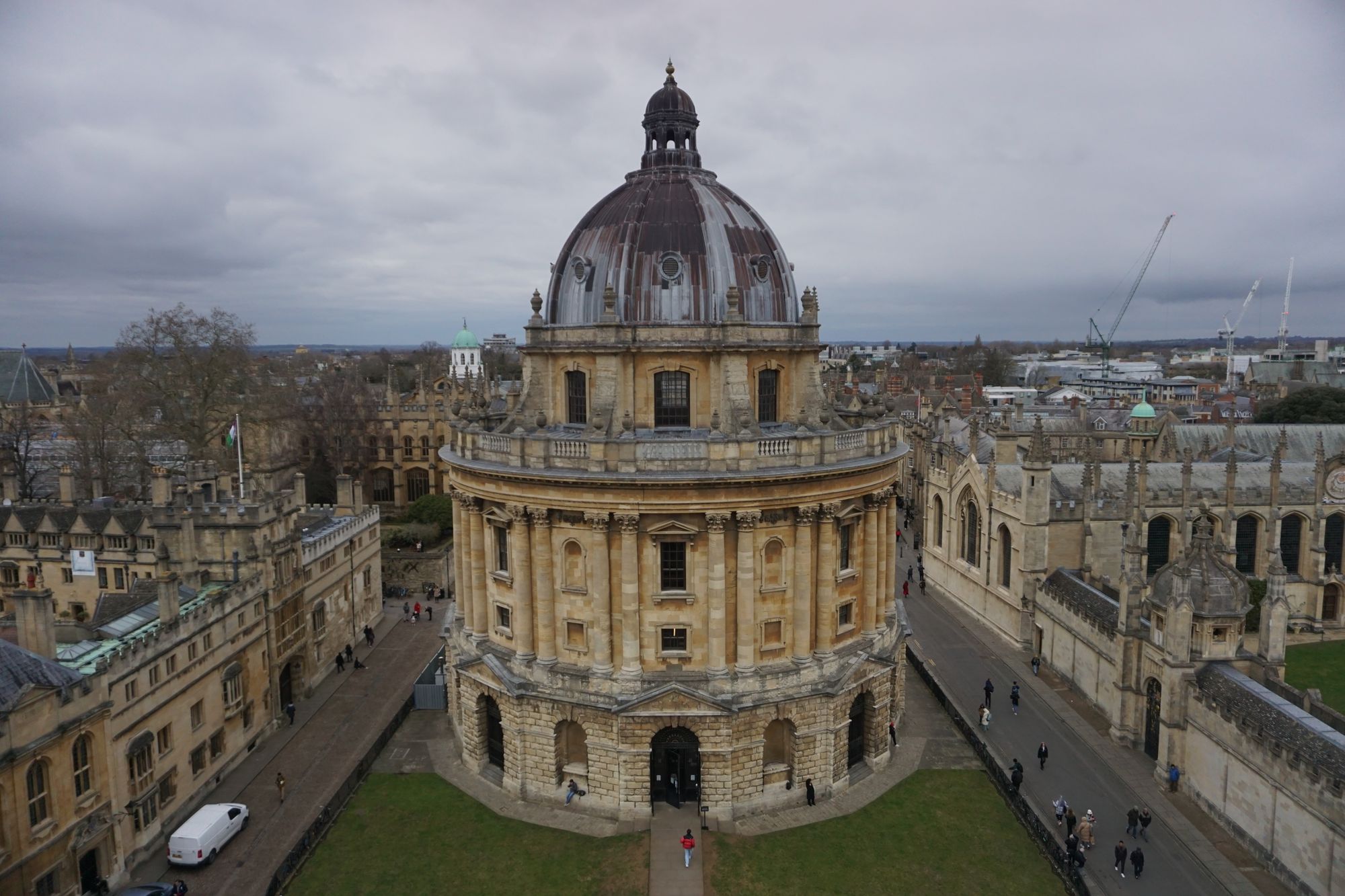 The Radcliffe Camera, an old circular stone building with a dome, in the centre of a pedestrianised square with drab greenery around it. A person in a red jacket walks towards the entrance.