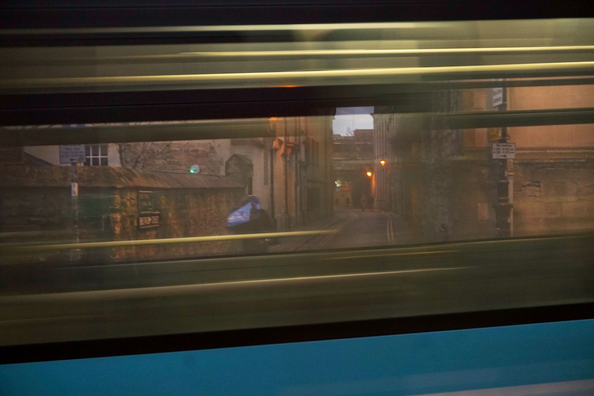 Seen through the blurred window of a passing bus, a cyclist turns down a narrow street with old buildings on either side.