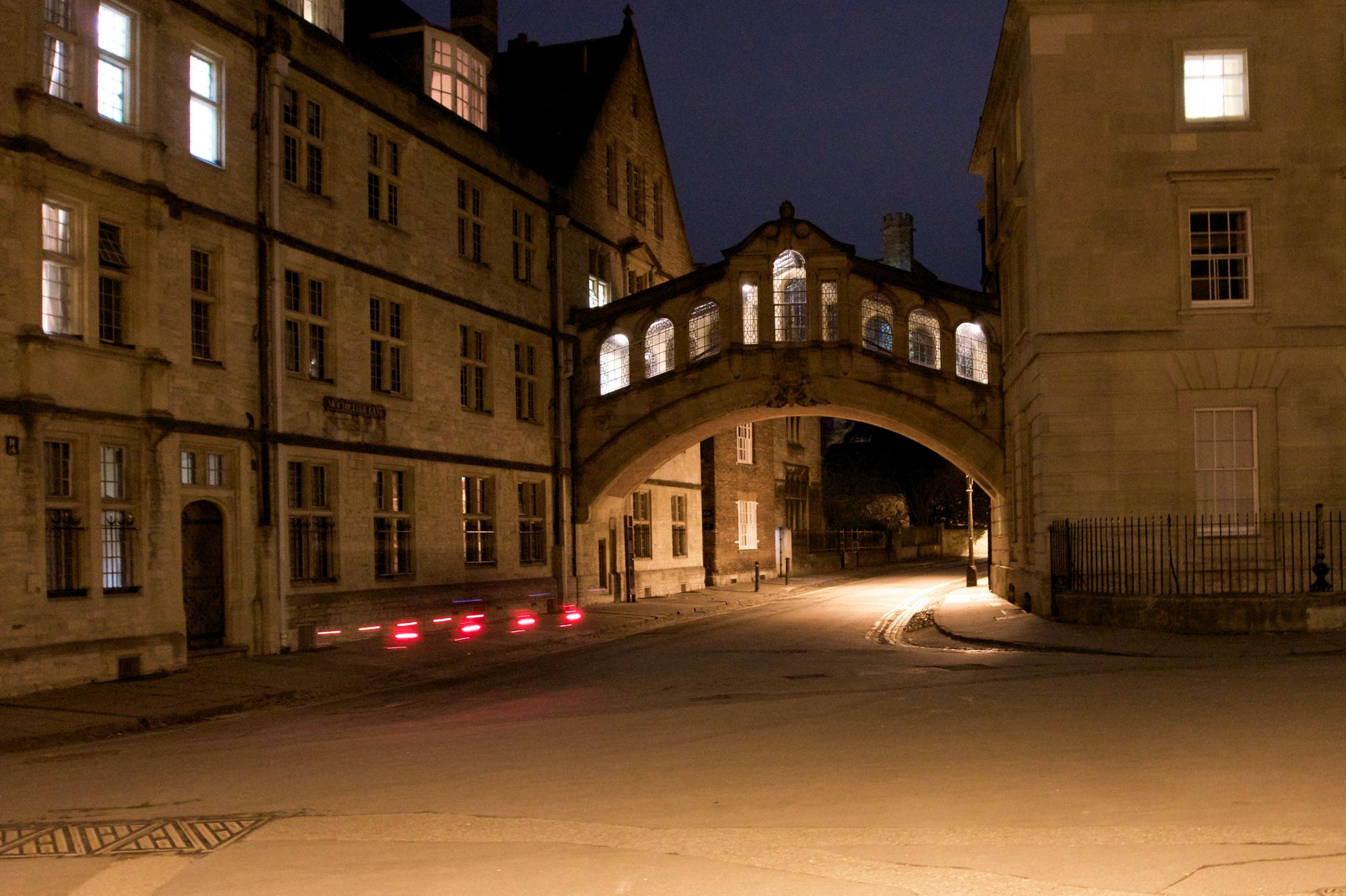 An arched bridge between two buildings over a street at night. The street is deserted save for some rear flashing cycle lights.