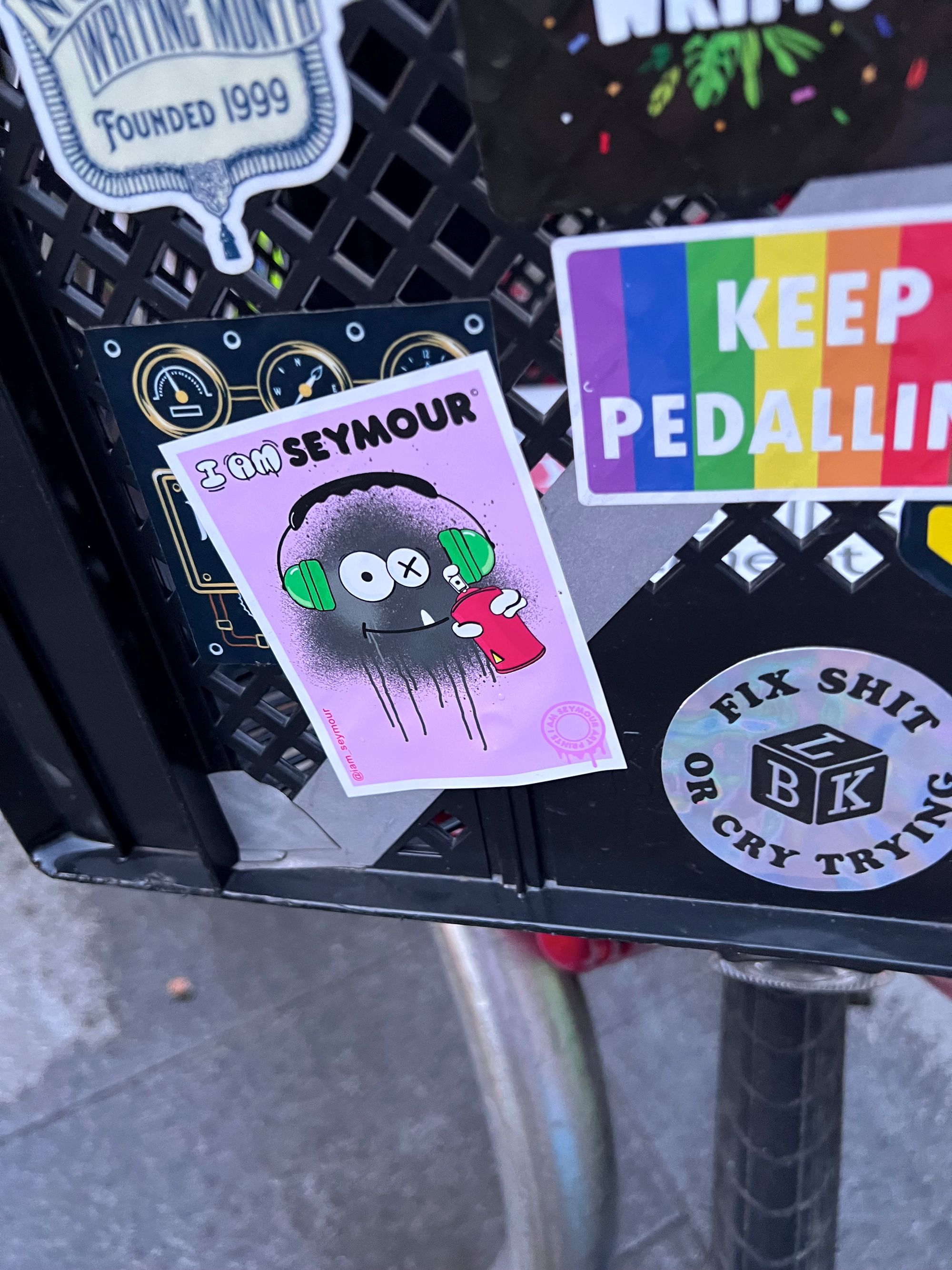 Close-up of the bicycle crate with a bevy of stickers, including one rectangular one - pink with a grey spray-painted face, holding a spray can and wearing headphones with one eye crossed out. It says "I AM SEYMOUR" and links to an apparent social media handle.