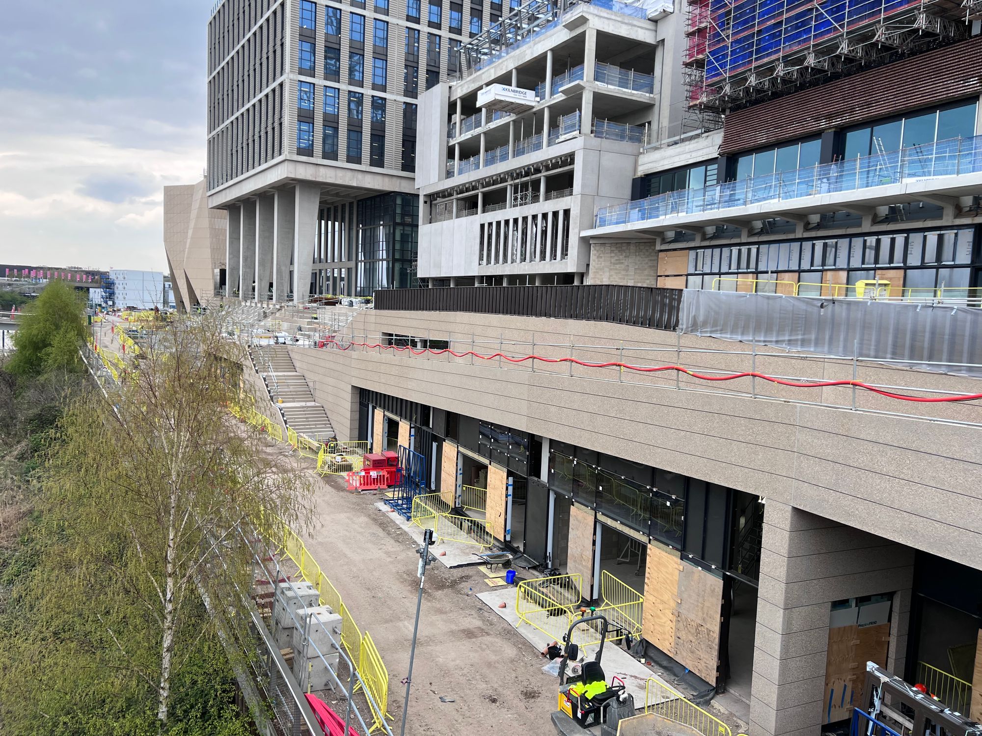 An under construction riverside development consisting of a river towpath, shop frontages, and stairs/lift arrangements leading up multiple levels. The cladding appears to be concrete.