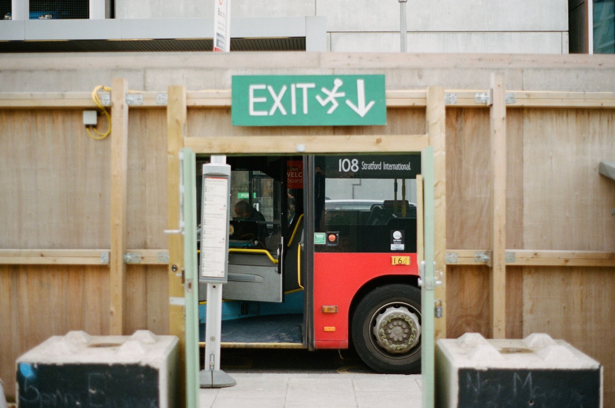 Looking through a wooden fence towards a bus standing at a stop with its doors open. It's a 108 to Stratford International. The driver is looking at something in his cab, and the door is open.