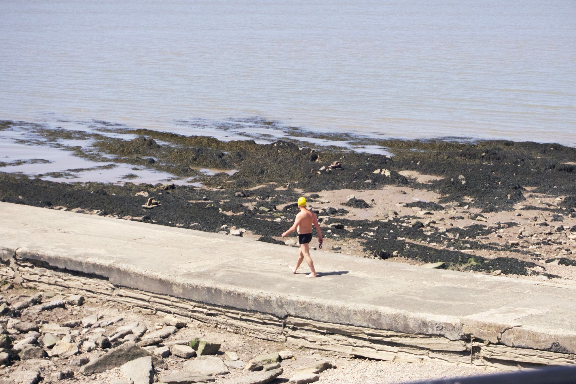 A swimmer in short black shorts and a yellow cap walks out along a groyne towards the Bristol Channel, low rock formations jutting out into the sea.