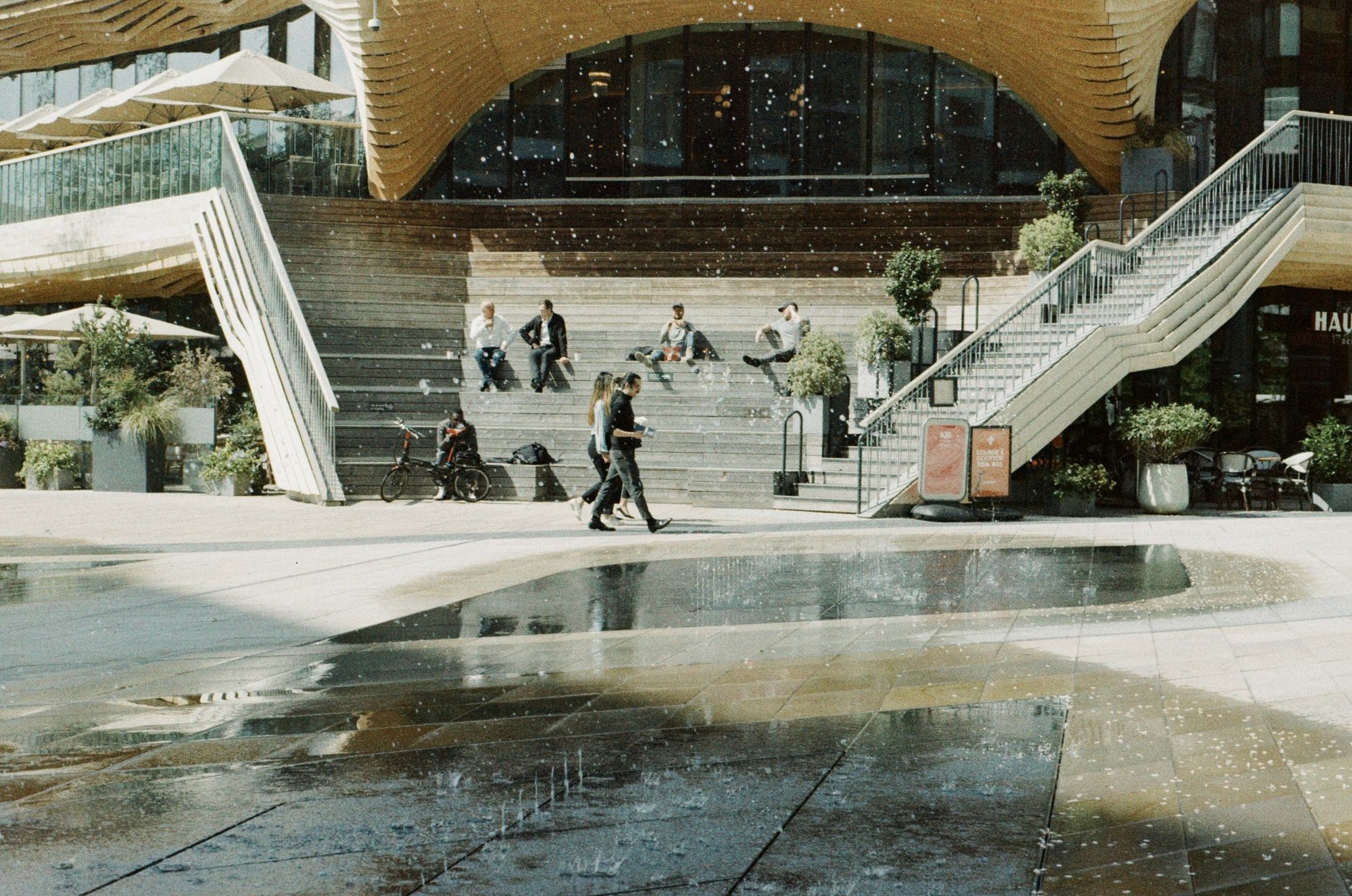 People sat on a wide staircase on a mock-wood effect building (the Haugen restaurant in Stratford) with digital fountains pulsing in the foreground. It’s a sunny day and there are sharp shadows on a mostly yellowy colour palette.