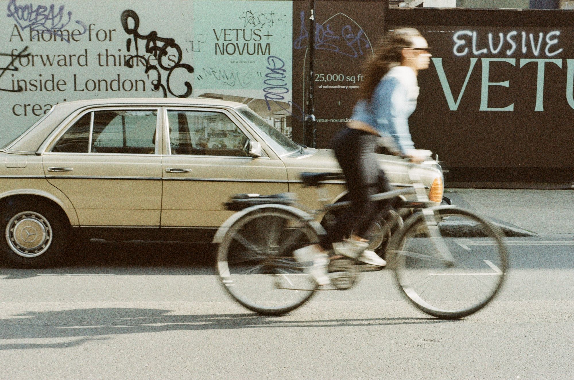 A woman in a blue top and black trousers with long, curly dark hair and sunglasses rides her bike past an old ochre/champagne coloured Mercedes parked next to some hoardings covered in graffiti. The colours are greyish and khaki with some strong contrast, and some colour coming from the woman’s top and the car’s indicator lenses.