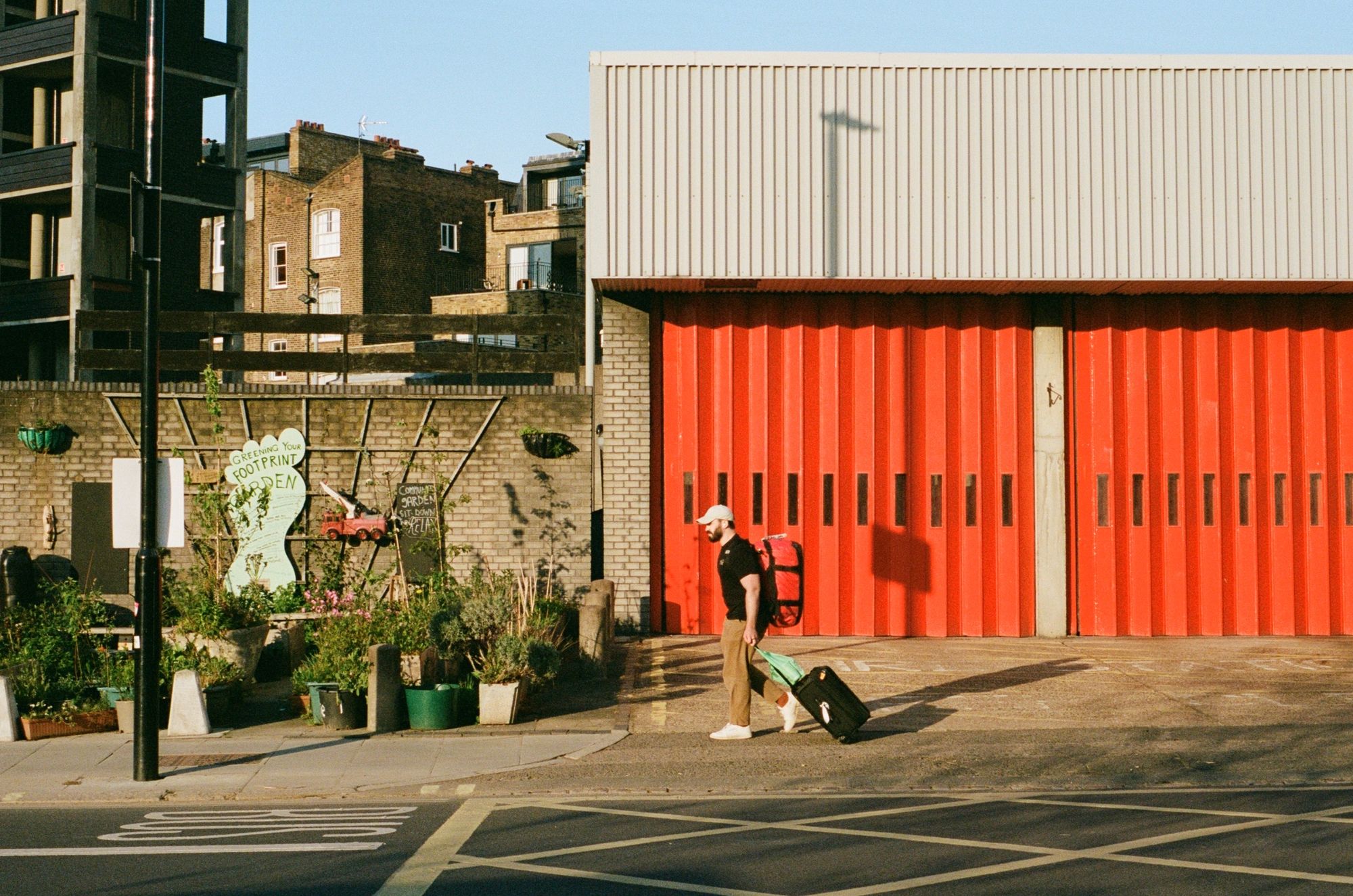 A man walks right to left across a pavement past a fire station on a sunny day, his black t-shirt contrasting against the red concertina doors. He has a wheely suitcase and a pale-coloured cap as he walks towards a community garden which is sited outside with a trellis on the wall and pots on the inner part of the pavement.