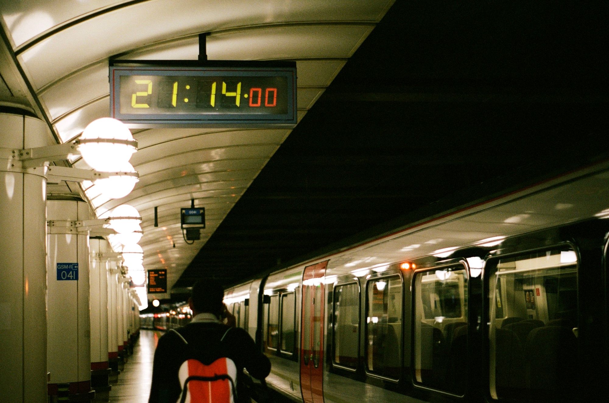 A man walks past a train with its doors unlocked but not open on a station platform lit by globe lights. The mechanical seven-segment clock reads 21:14:00.