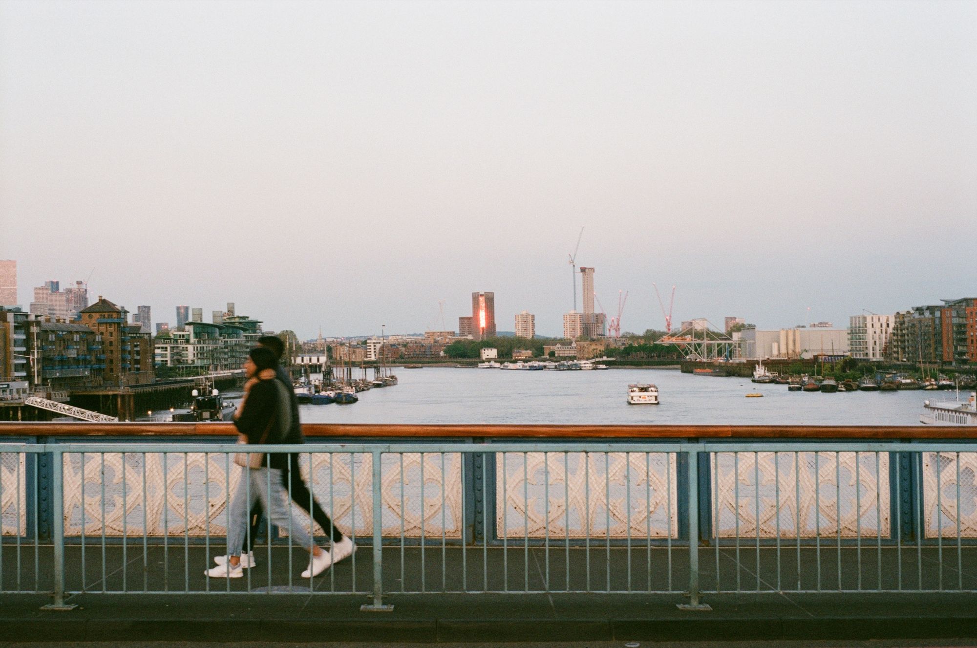 Looking east from Tower Bridge over the River Thames, we see people walking right to left in frame over the opposite walkway (between pedestrian guardrail painted pale blue and a thick, ornate balustrade painted dark blue and white with a wooden top.) In the distance, a block of apartments has caught the light of the setting sun shining a thin strip of orange reflection.