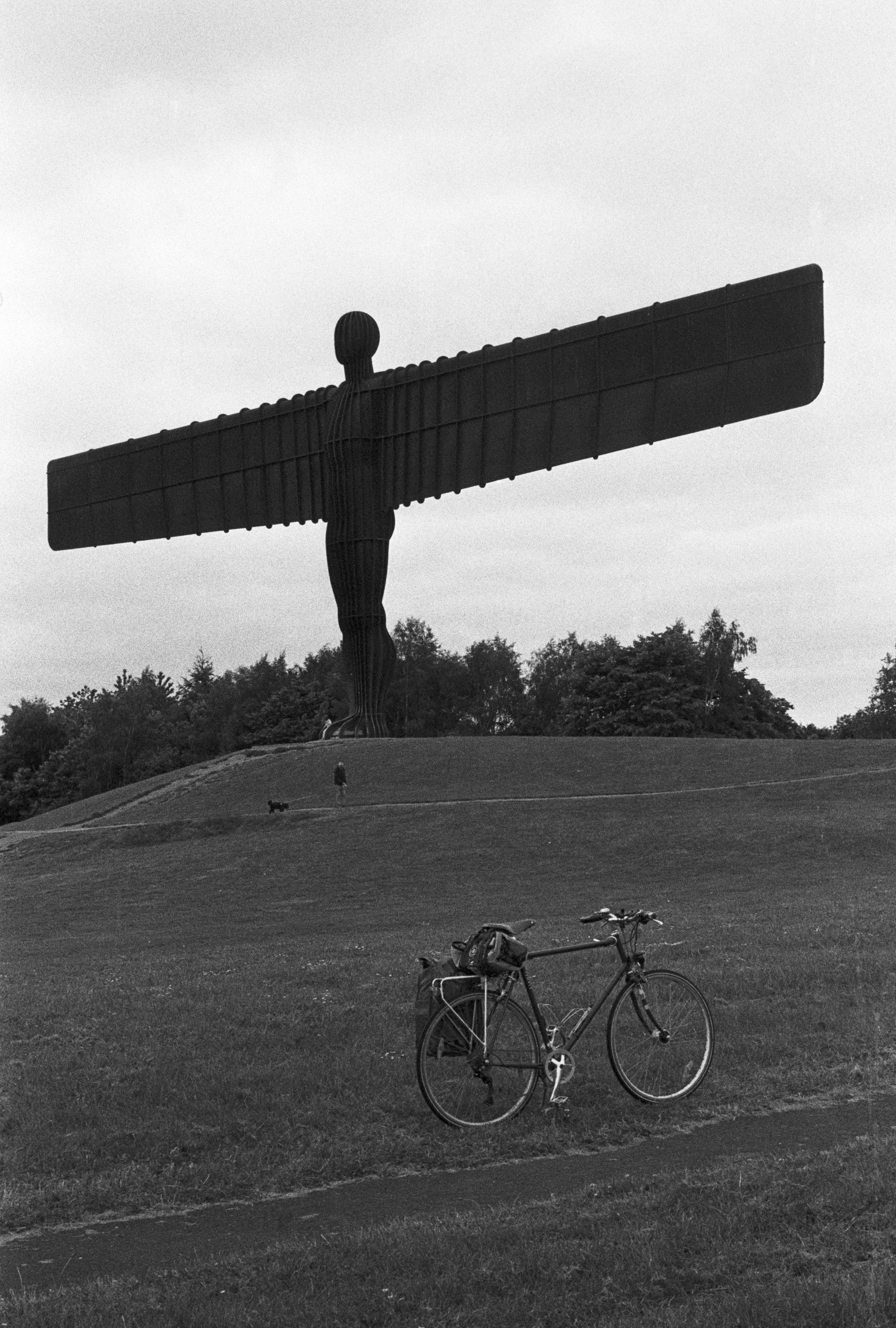 Jonathan's bike in front of the Angel of the North, a large metal statue in the shape of a person standing with aeroplane wings, in black and white.