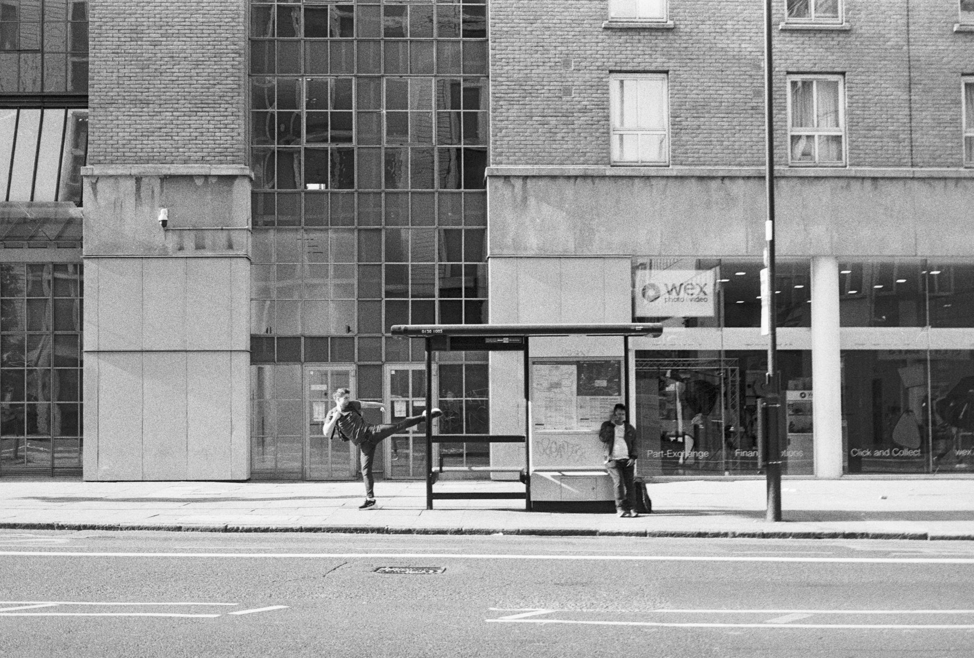 A bus stop on a large road outside a glass shop called "Wex Photo Video." A man sits in the shelter on the phone. Another man does mock martial arts including a karate kick on the pavement, just behind the bus shelter.