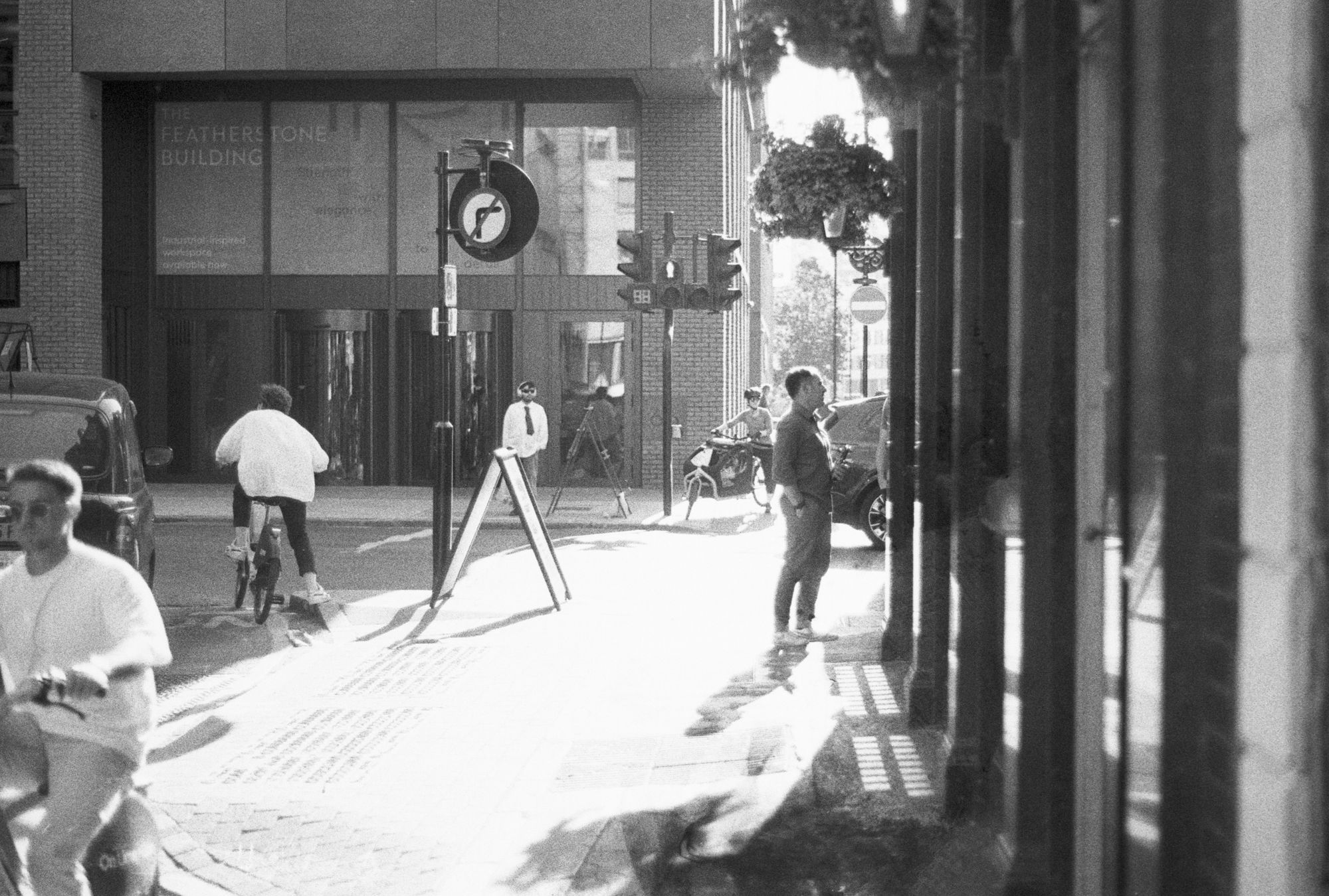 The junction of a street with a major road with another street in the distance, with harsh contrast lighting. A taxi and a cyclist on a Lime bike leave the street. Another cyclist enters the street. Another cyclist waits to cross the main road on the shared crossing. A man stands outside a pub casting a shadow. An A-board stands parallel to the kerb line next to a no-right-turn signpost probably getting in the way. All rendered in black and white with blown-out highlights and soft shadows.