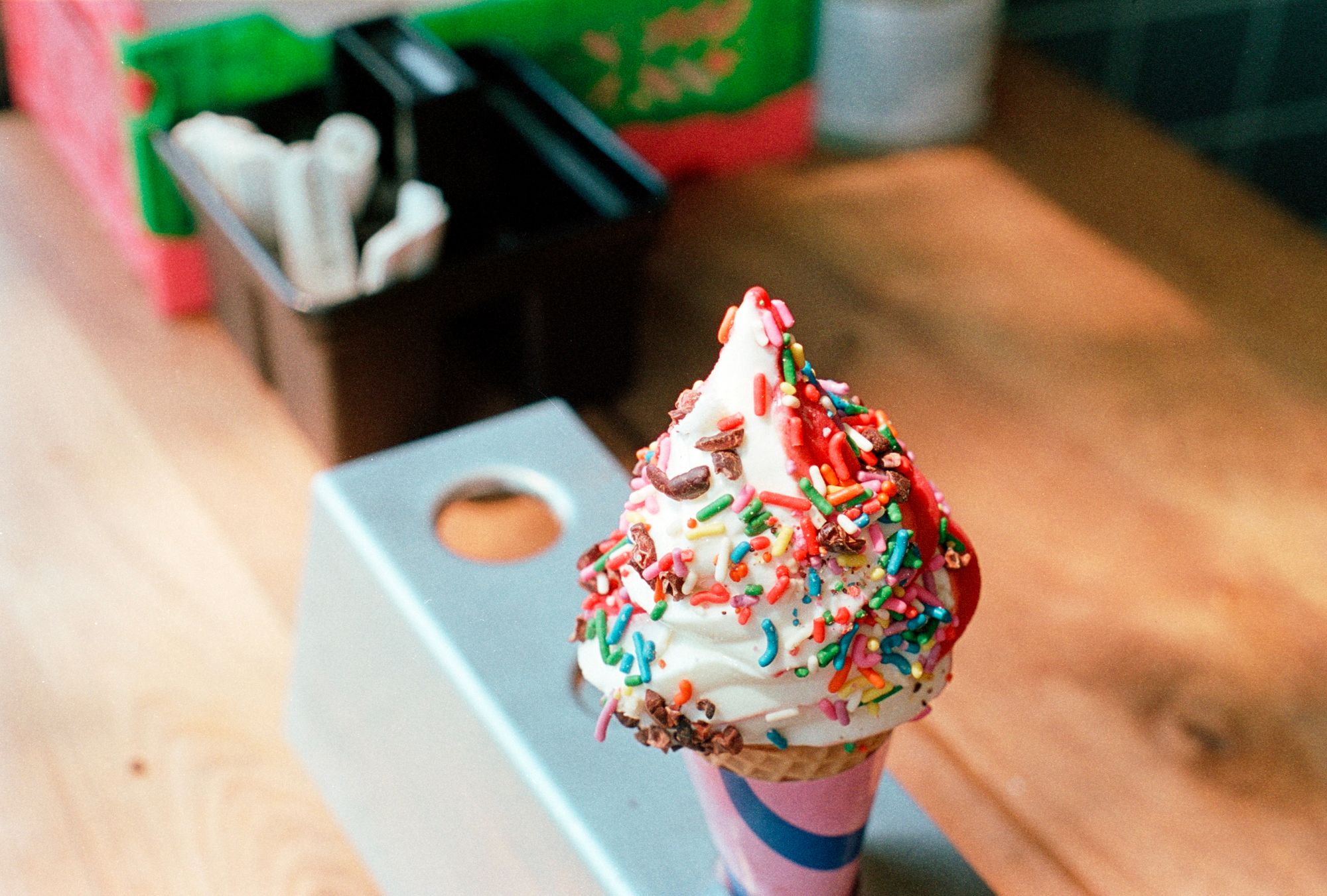 An ice cream cone with colourful sprinkles, partly vanilla and partly raspberry sorbet, in a holder on a wooden counter.
