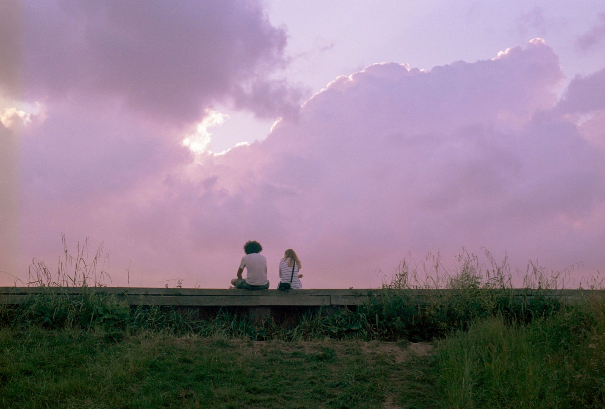 Against a muted pink-purple sunset with fluffy cirrus clouds forming a silver lining behind their tempestuous mauve colours: two people in casual clothes sit on a low wooden wall, facing away from camera, at the top of a grassy ridge.