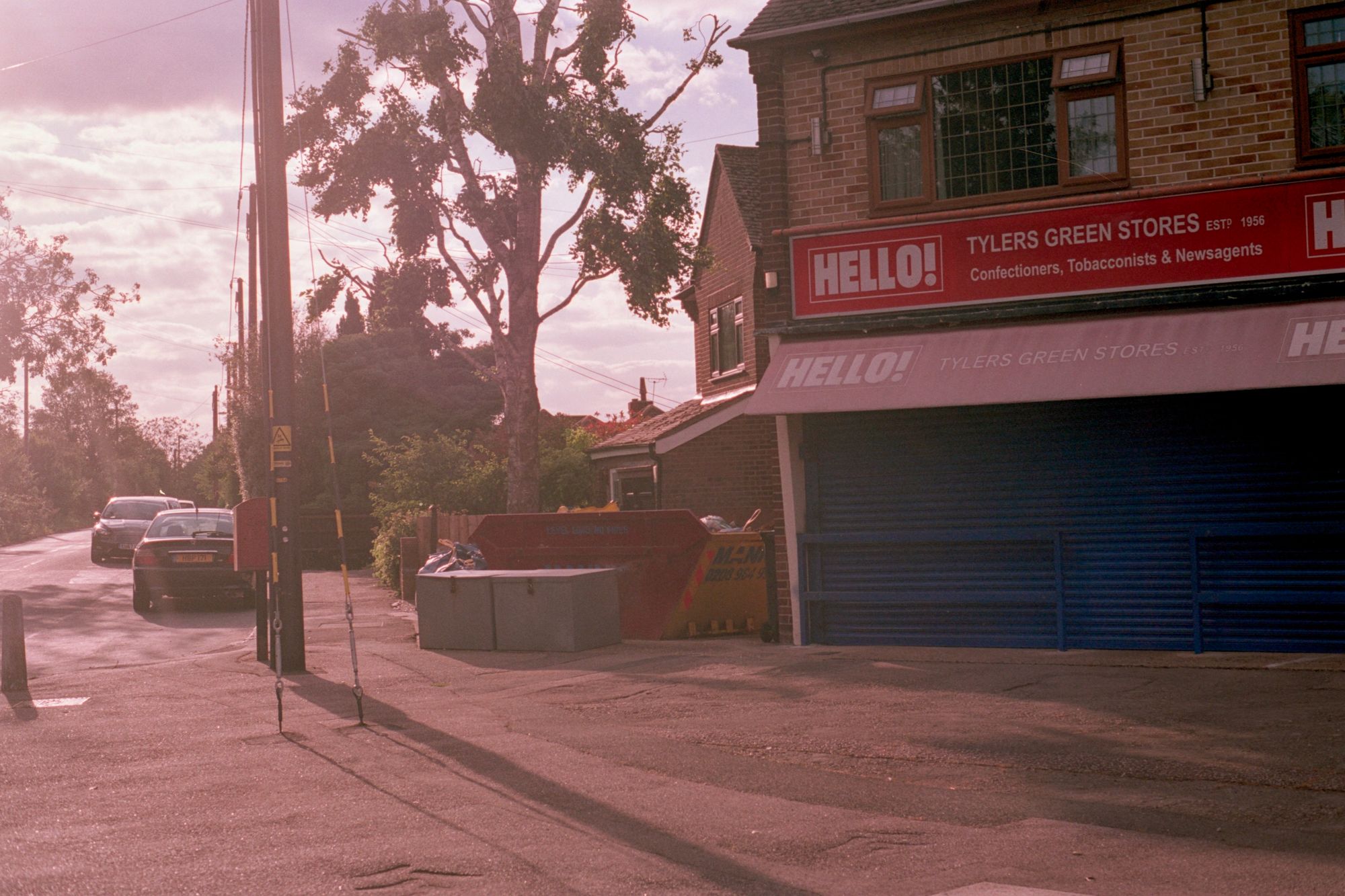 At sunset, A corner shop with its shutters closed: "Tylers Green Stores, established 1956, confectioners, tobacconists & newsagents." The logo of HELLO! magazine appears ton the shop frontage and the faded red awning. There's a telegraph pole and a skip.
